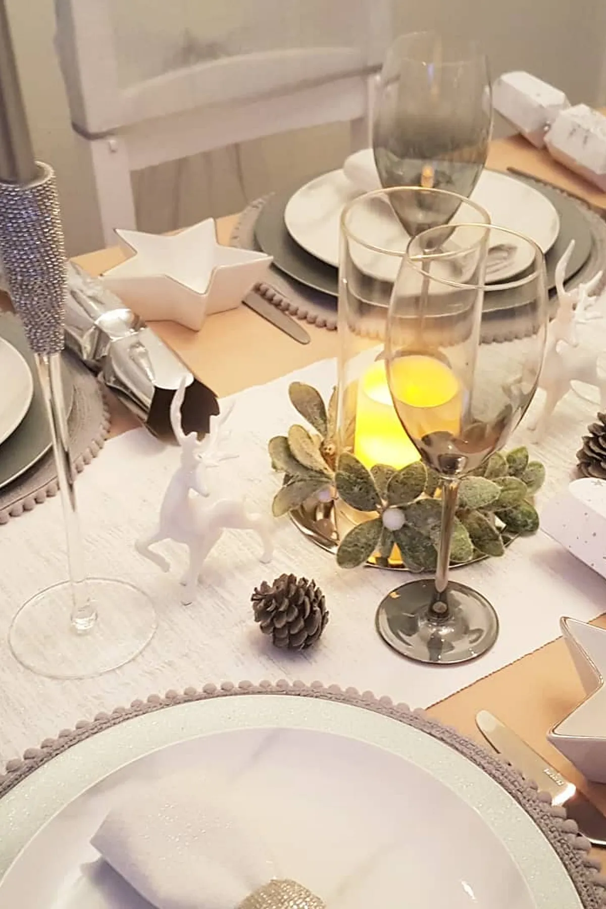 Festively set dining table with white plates, silver utensils, a candlestick, reindeer figurines, and star-shaped dishes on a warm wooden surface.