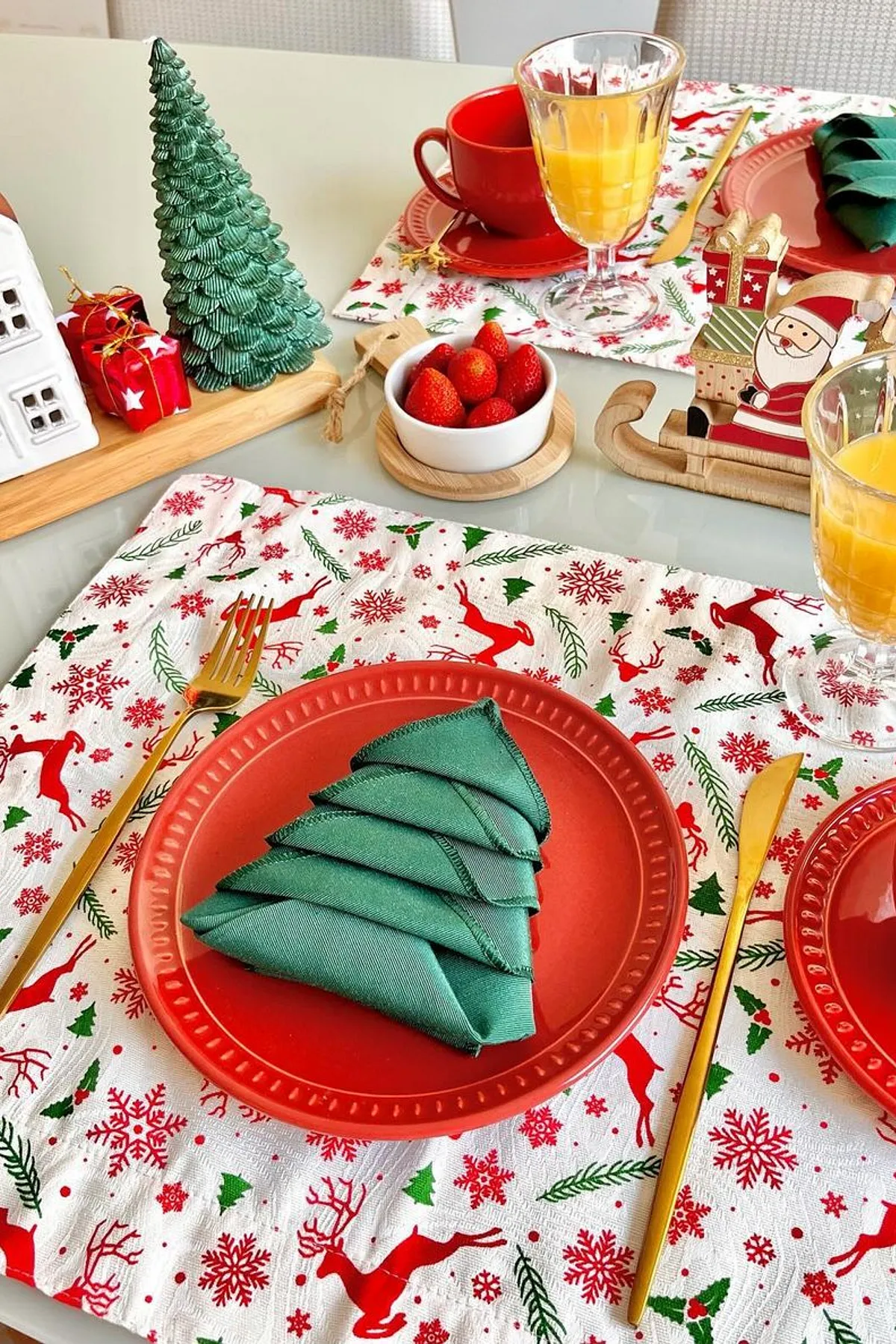 Festive Christmas table setup with red placemats, tree-shaped napkin, golden cutlery, wooden house decor, and a bowl of strawberries.