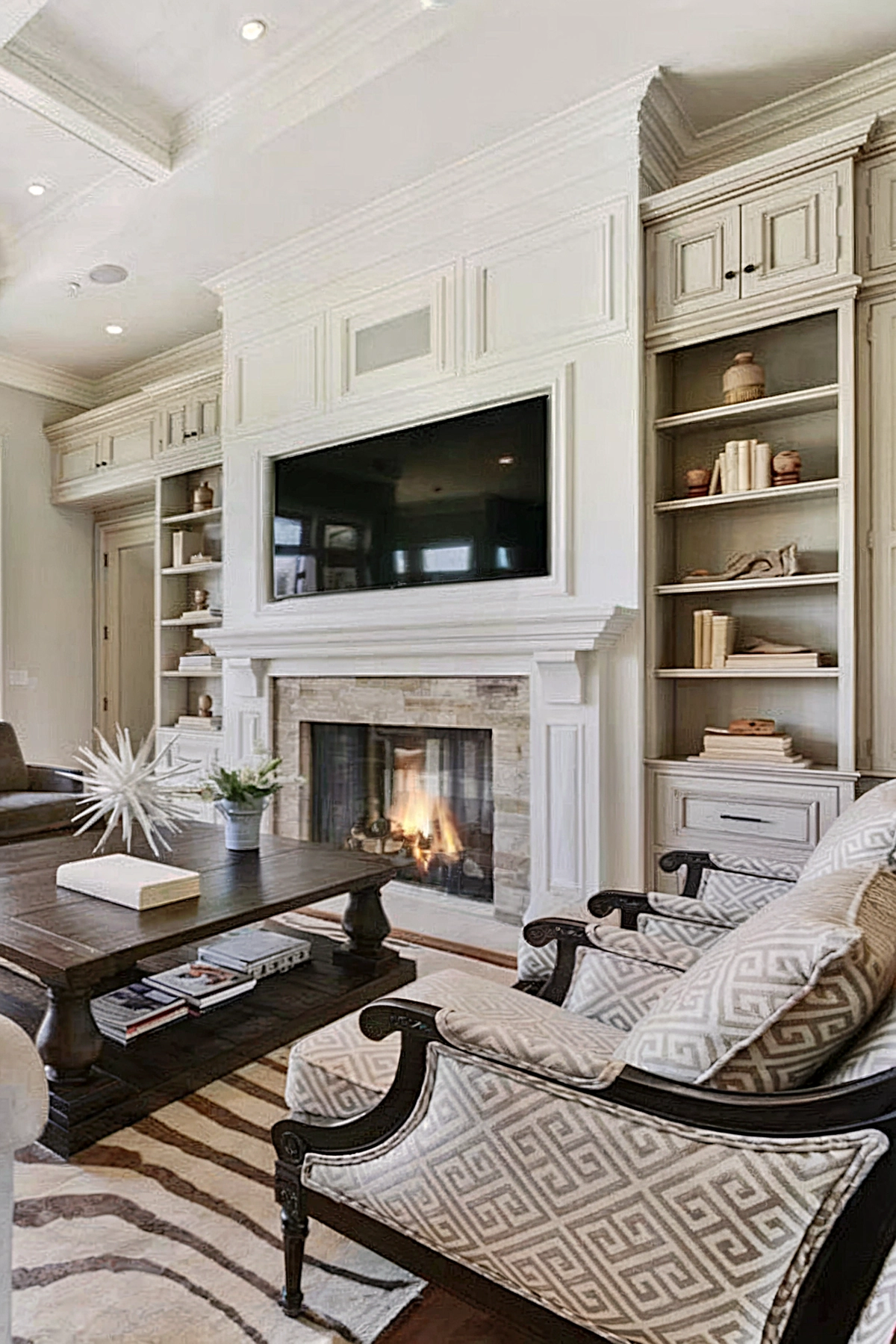 A well-lit living room with a prominent fireplace flanked by built-in shelves, a large TV above the mantel, and patterned armchairs surrounding a dark wood coffee table