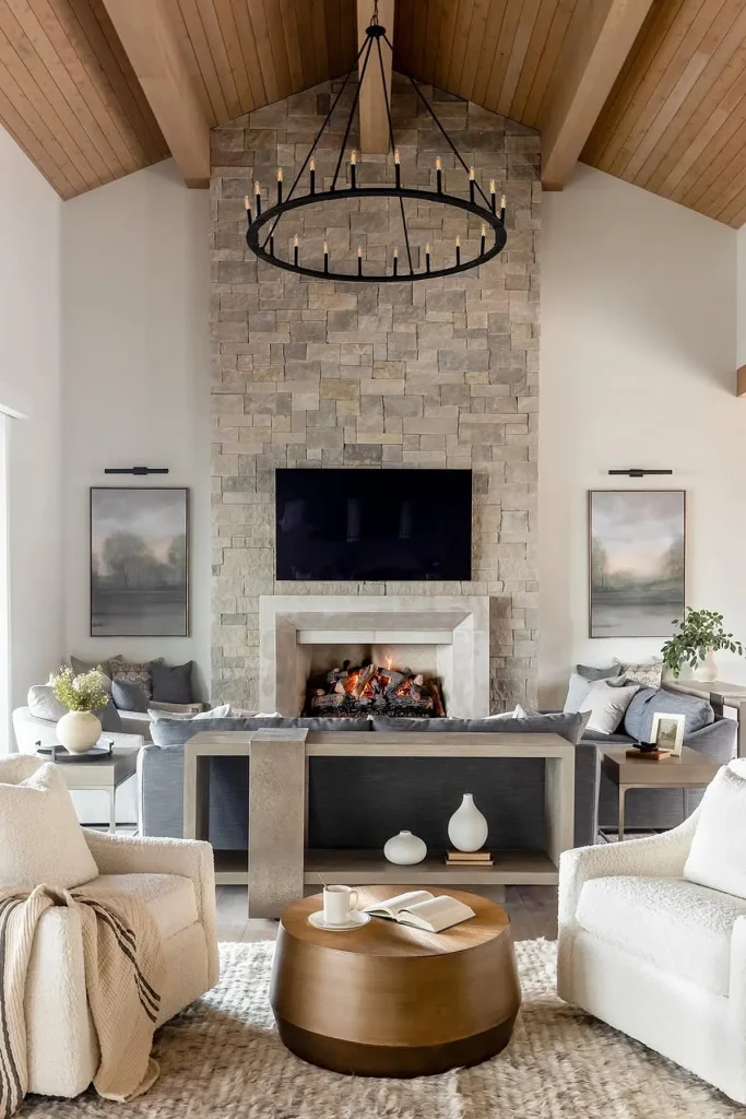A modern rustic living room featuring a stone fireplace with a black circular chandelier hanging above. Two cozy white armchairs and a round wooden coffee table sit on a textured rug, surrounded by neutral-toned decor and soft furnishings. Natural wood beams and framed artwork complete the serene space