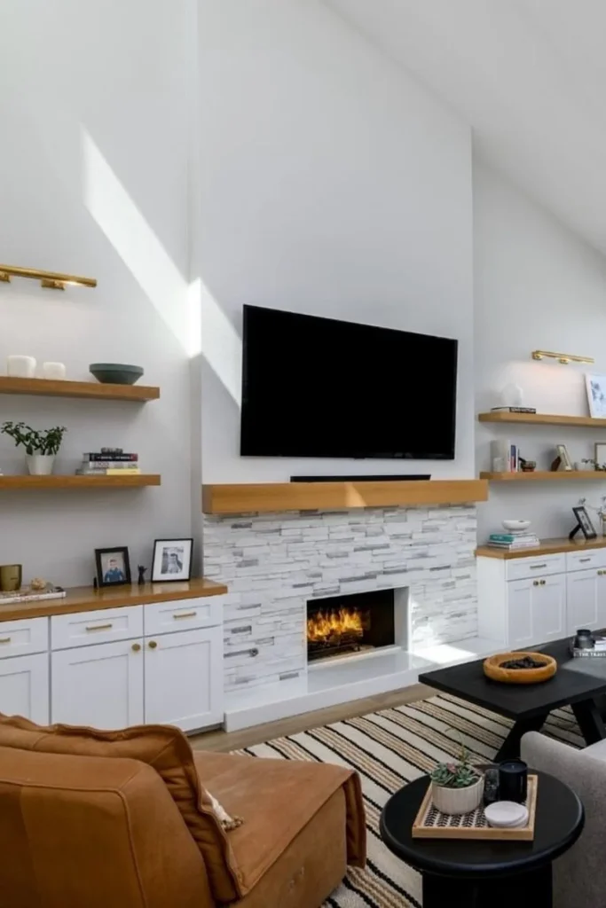 A modern living room with a TV mounted above a white stone fireplace, featuring wooden floating shelves and built-in cabinets. A tan leather chair, striped rug, and black coffee table complete the space, adding warmth and texture