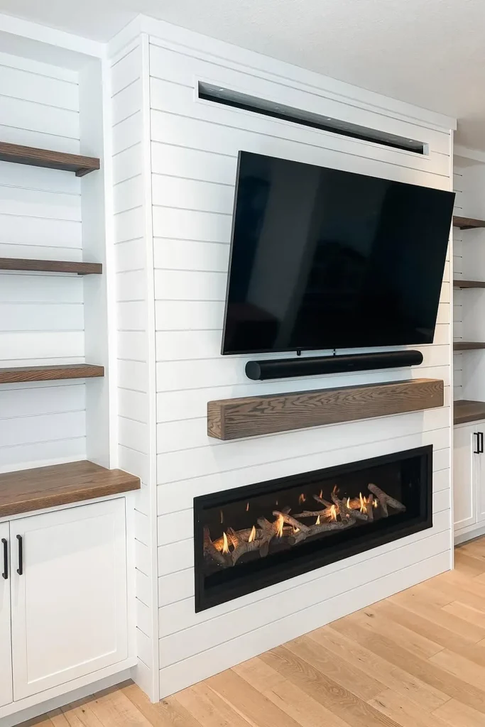 A modern living room featuring a white shiplap fireplace wall with a mounted TV above a floating wood mantel. A sleek gas fireplace adds warmth, while built-in cabinets and open wood shelves provide storage and style