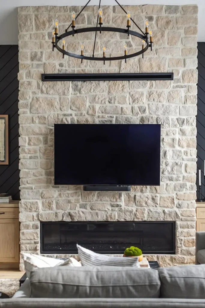 living room featuring a stone fireplace wall with a mounted TV and a linear gas fireplace below. A black circular chandelier hangs above, and dark wood accents frame the space, creating a blend of rustic and modern design