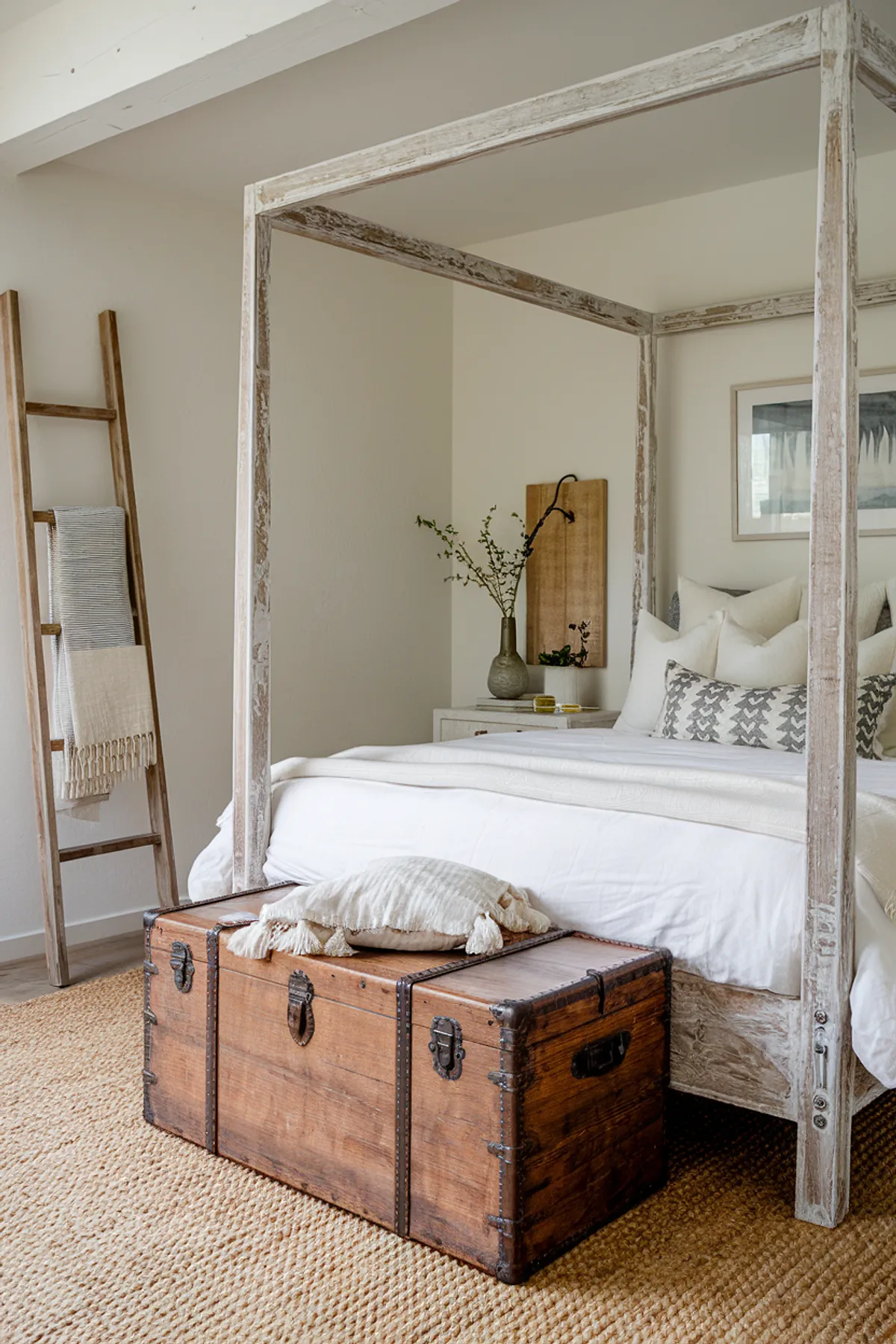 Bedroom with rustic canopy frame, white linens, wooden trunk, and framed artwork.