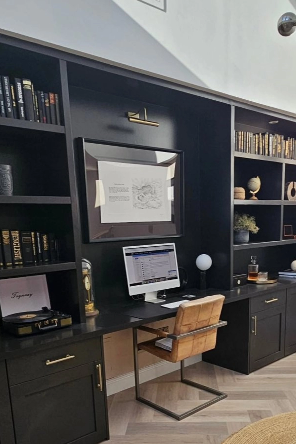A sophisticated home office with black built-in shelving, a computer workstation, warm leather chair, gold hardware, and a curated mix of books and decorative accents.