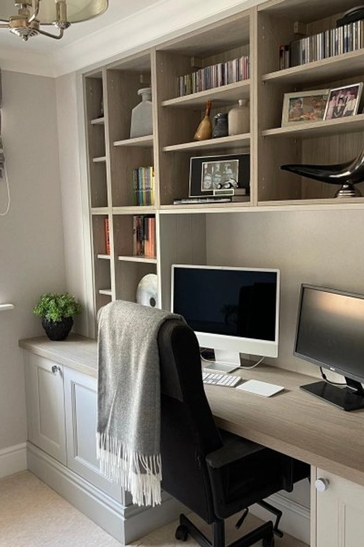 A home office setup with built-in light wood shelving, a computer workstation with dual monitors, a black office chair with a gray throw, and decor including books, framed photos, and vases.