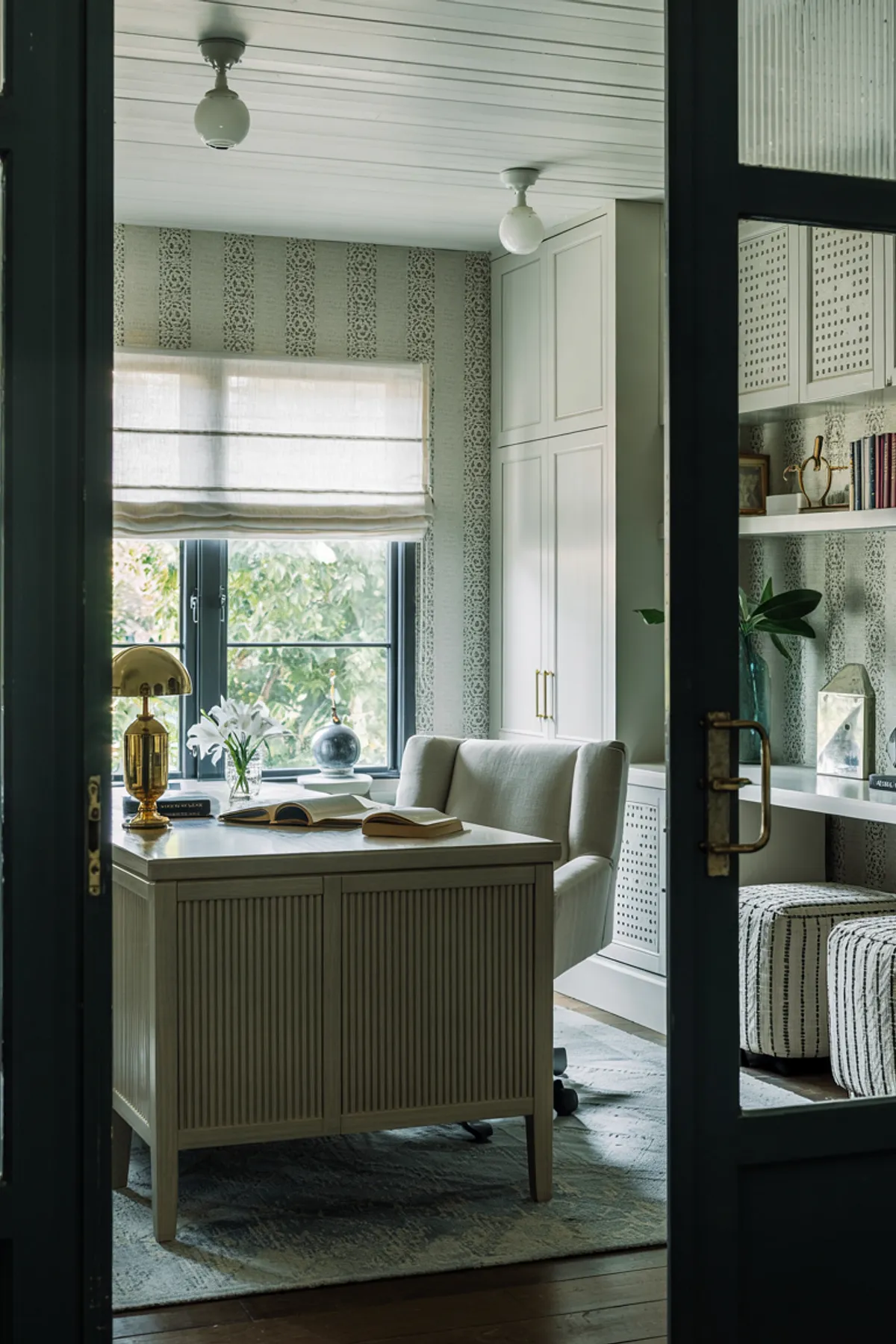 Home office with wooden desk, large window showcasing greenery, open shelving, and cozy decorative accents.