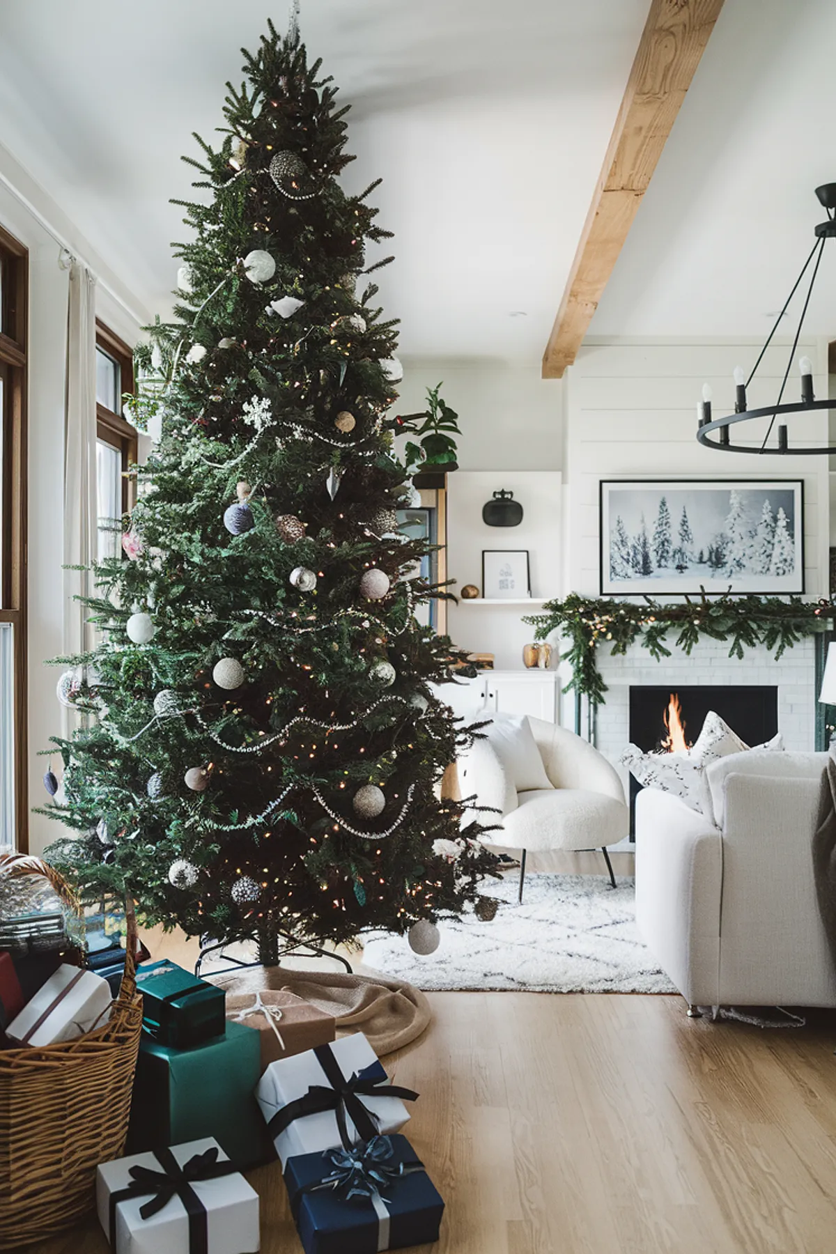 Cozy holiday living room with a decorated Christmas tree, fireplace, modern chandelier, and large windows letting in natural light.