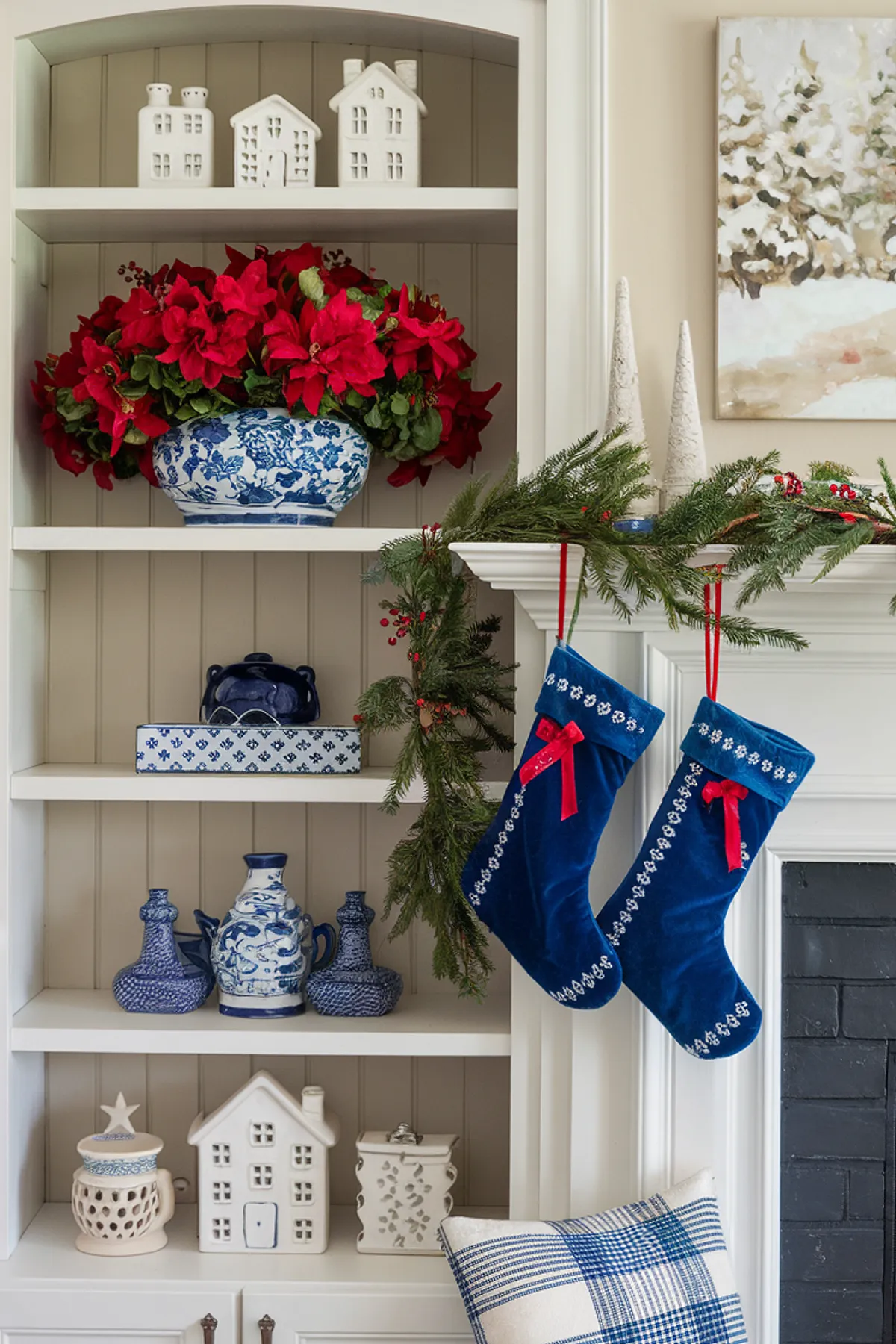 White shelving unit decorated with blue and white porcelain, red poinsettias, and a white fireplace mantle adorned with greenery and stockings.