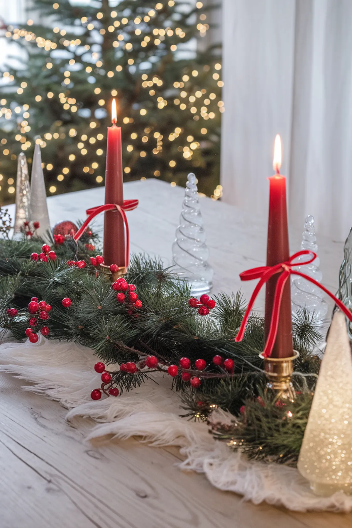 Festive Christmas table setting with pine garland, red candles, glass ornaments, and a beautifully lit Christmas tree in the background.