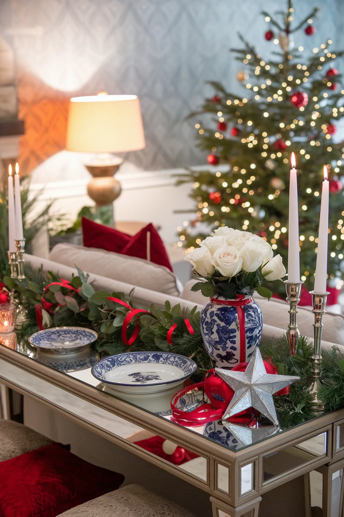 Cozy holiday living room with a Christmas tree, mirrored console table, white roses, candles, red accents, and warm lighting.