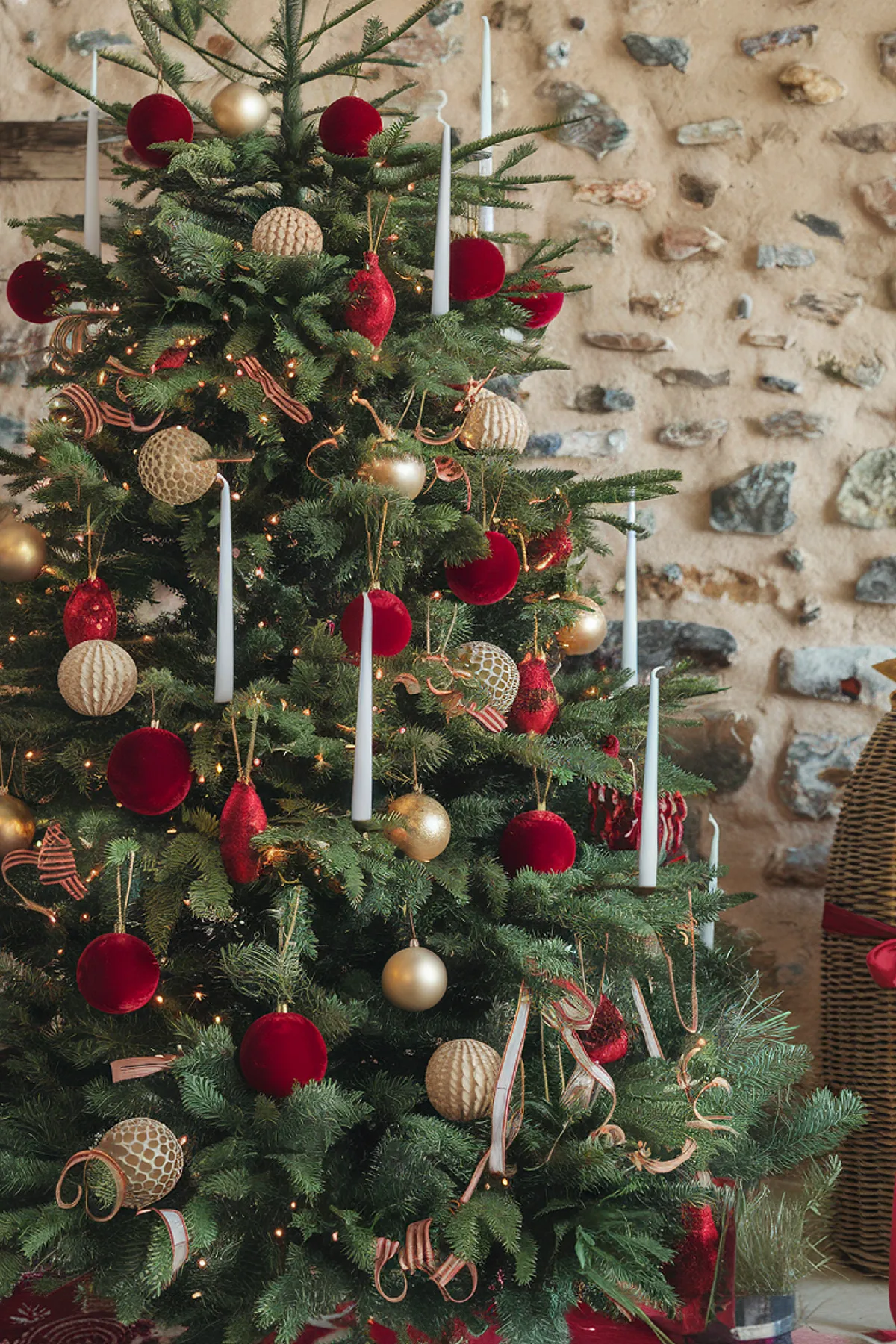 Decorated Christmas tree with red and gold ornaments, white candles, wicker basket, patterned rug, and wrapped gifts beneath a stone wall.