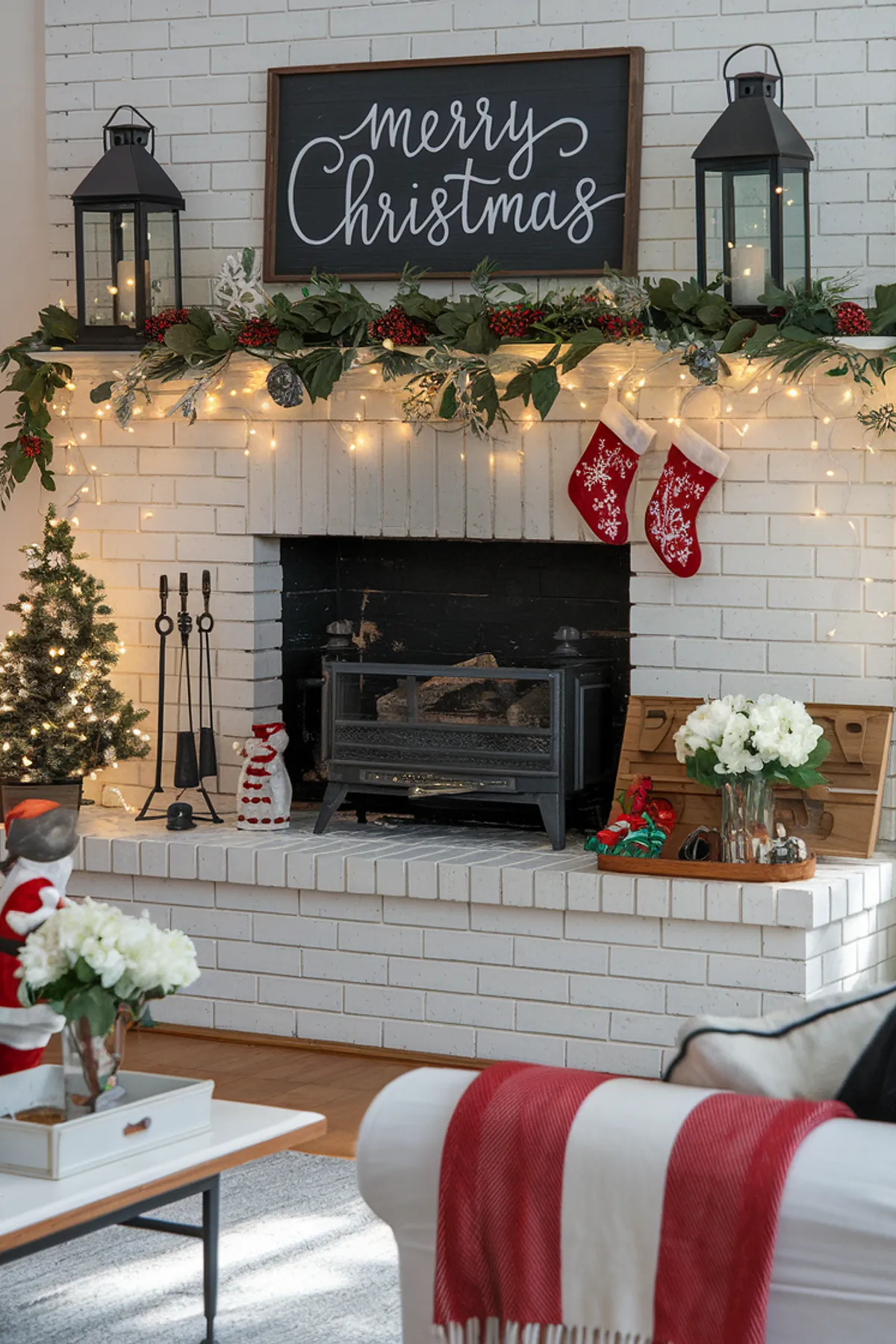 Cozy holiday living room with white brick fireplace, Christmas tree, stockings, lanterns, and a white couch adorned with a striped blanket.