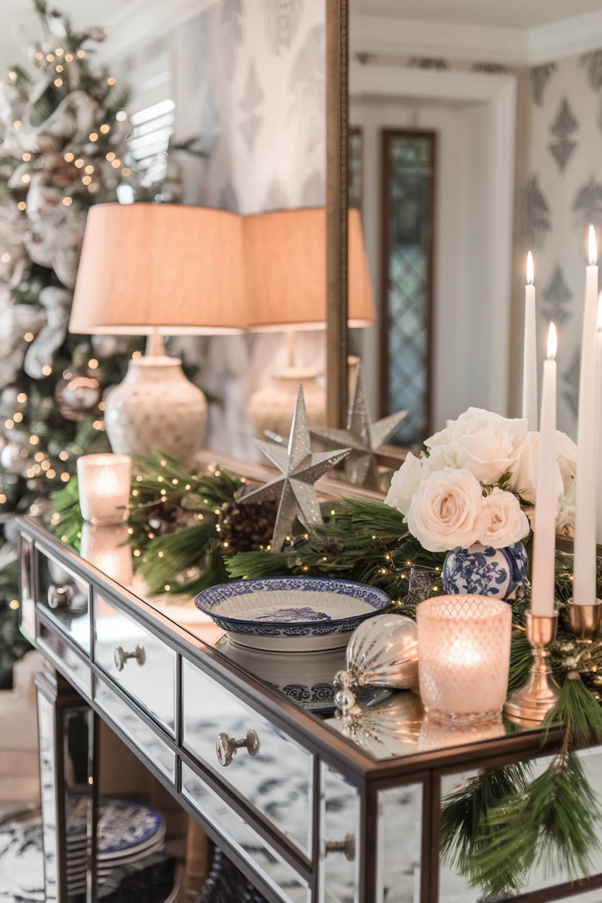 Elegant interior with a mirrored console, white roses, festive Christmas tree, and warm lighting in a stylishly decorated space.