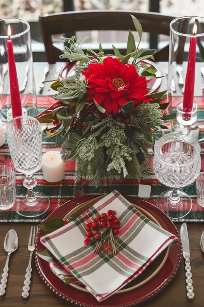 Festively set dining table with red flower centerpiece, plaid napkin, candles, and glassware against a window backdrop