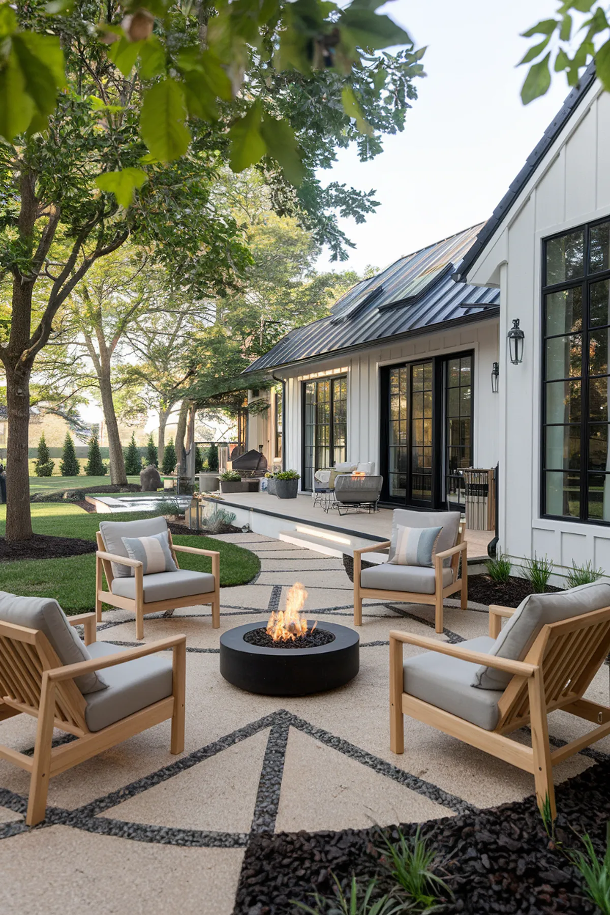 Modern patio with wooden chairs around a fire pit, surrounded by trees and a white house with dark roof.
