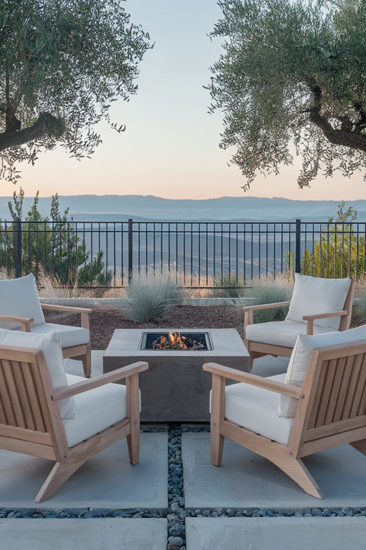Outdoor seating area with wooden chairs around a rectangular fire pit, flanked by olive trees and ornamental grasses.