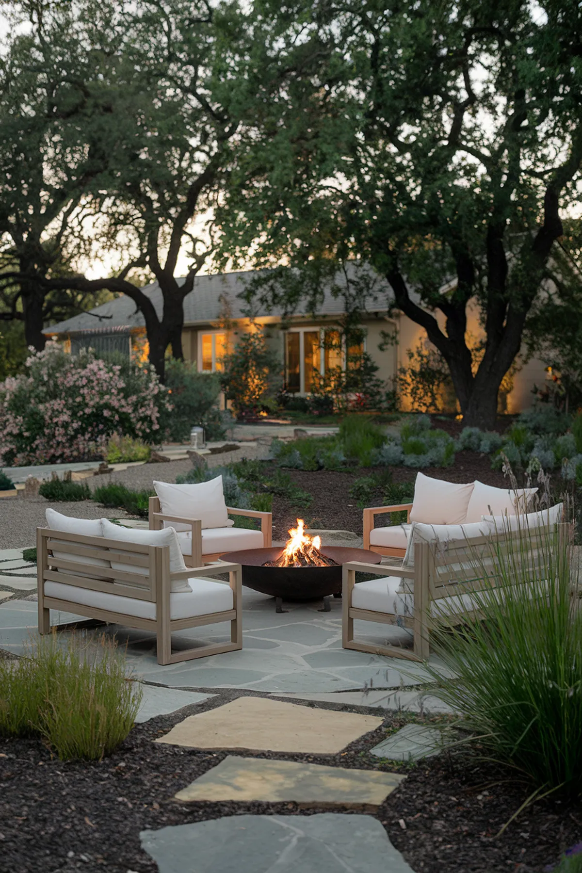 Evening outdoor seating area with wooden sofas and fire pit surrounded by trees and a house with warmly lit windows.