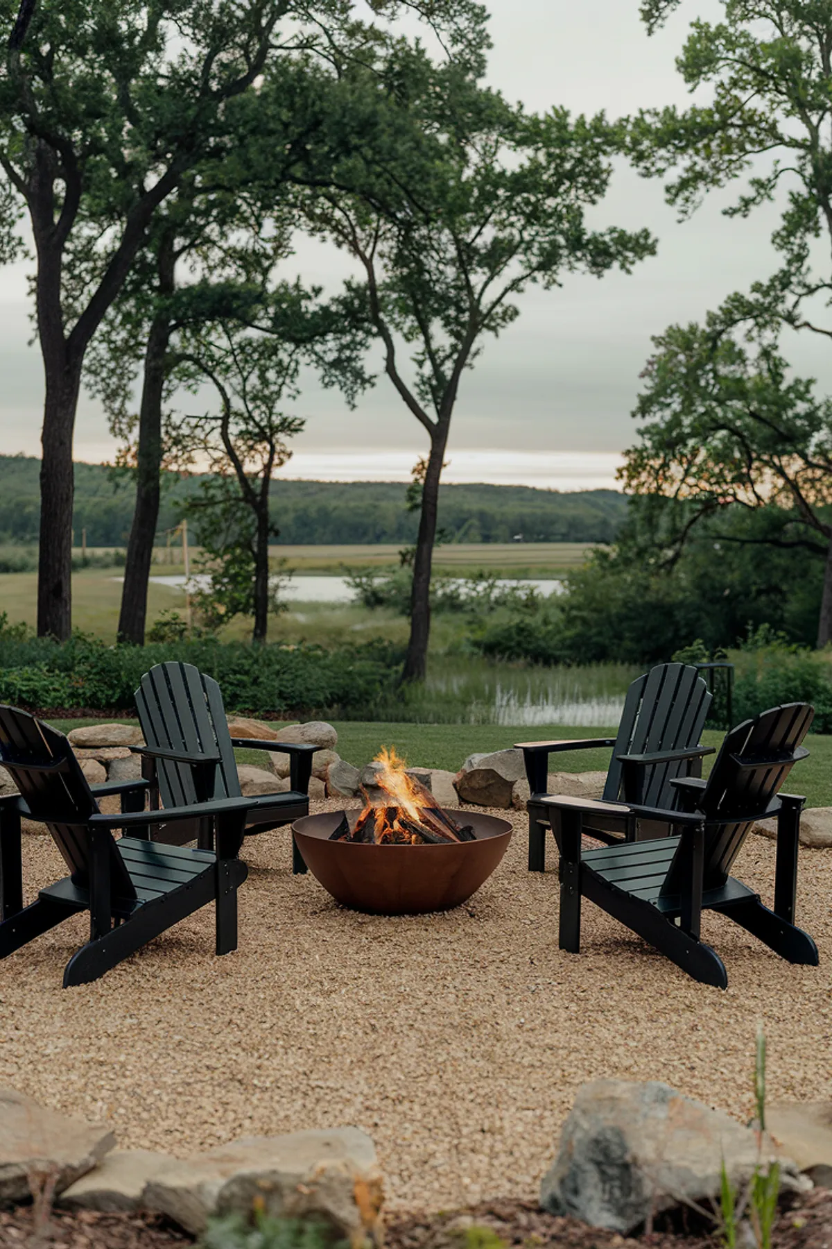 Outdoor dusk setting with a fire pit, black Adirondack chairs on gravel, and a view of a water body and hills.