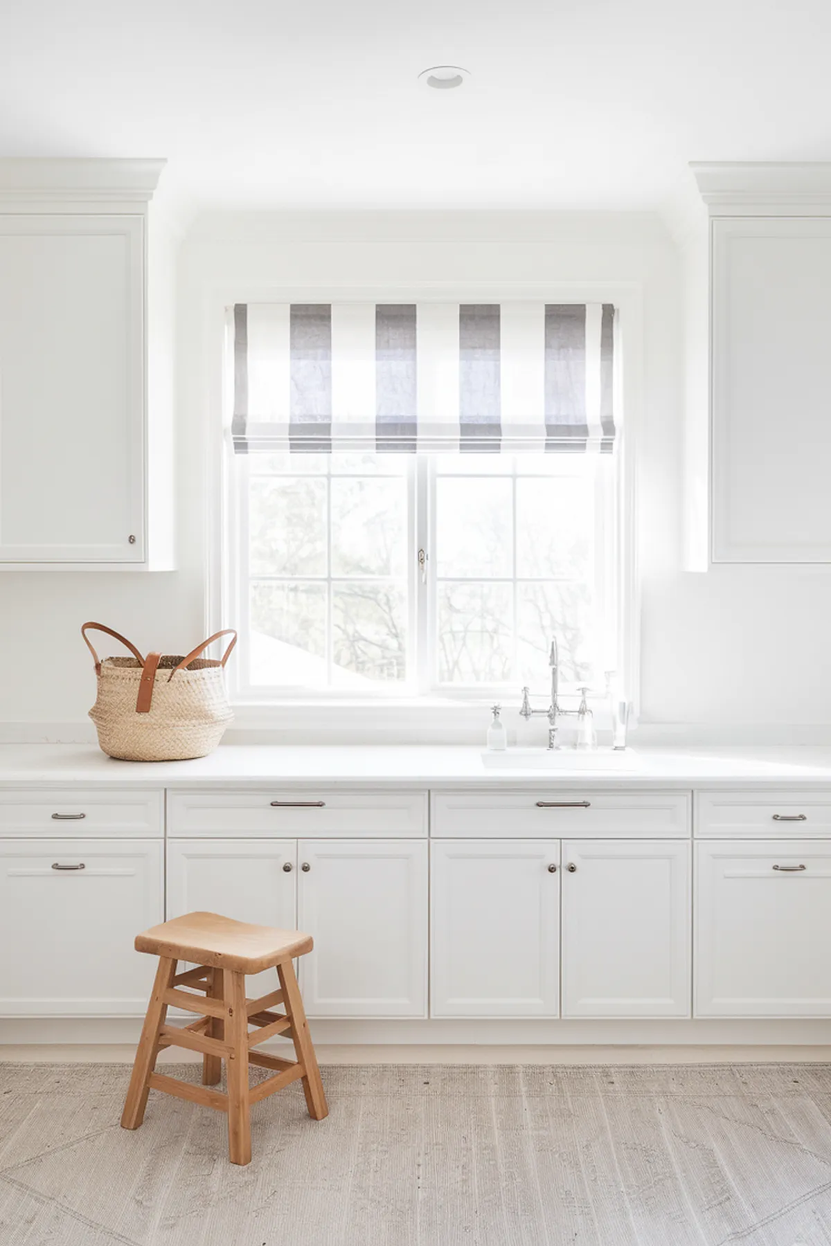 A bright laundry room featuring white cabinets, a striped window treatment, and a wooden stool.