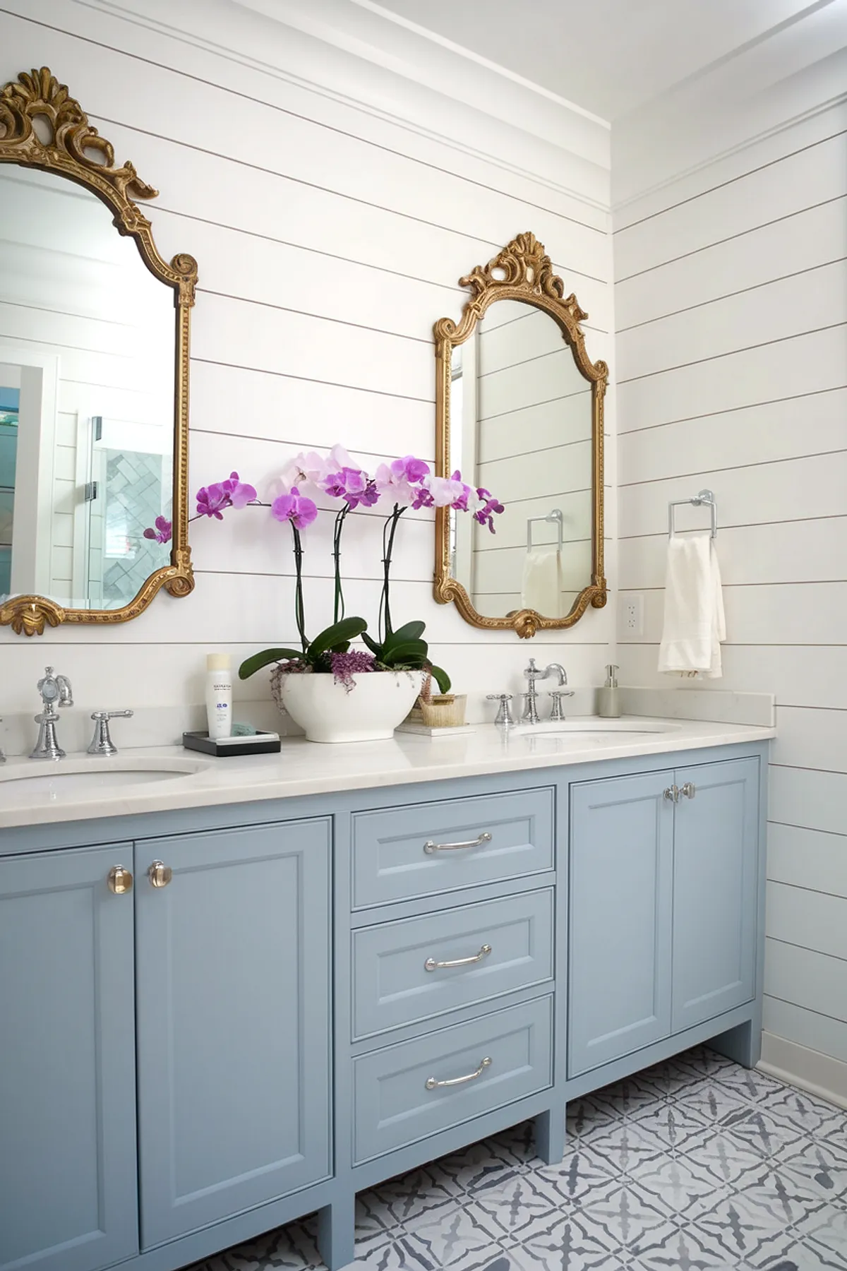 A stylish bathroom featuring blue vanity, gold-framed mirrors, and purple orchids.