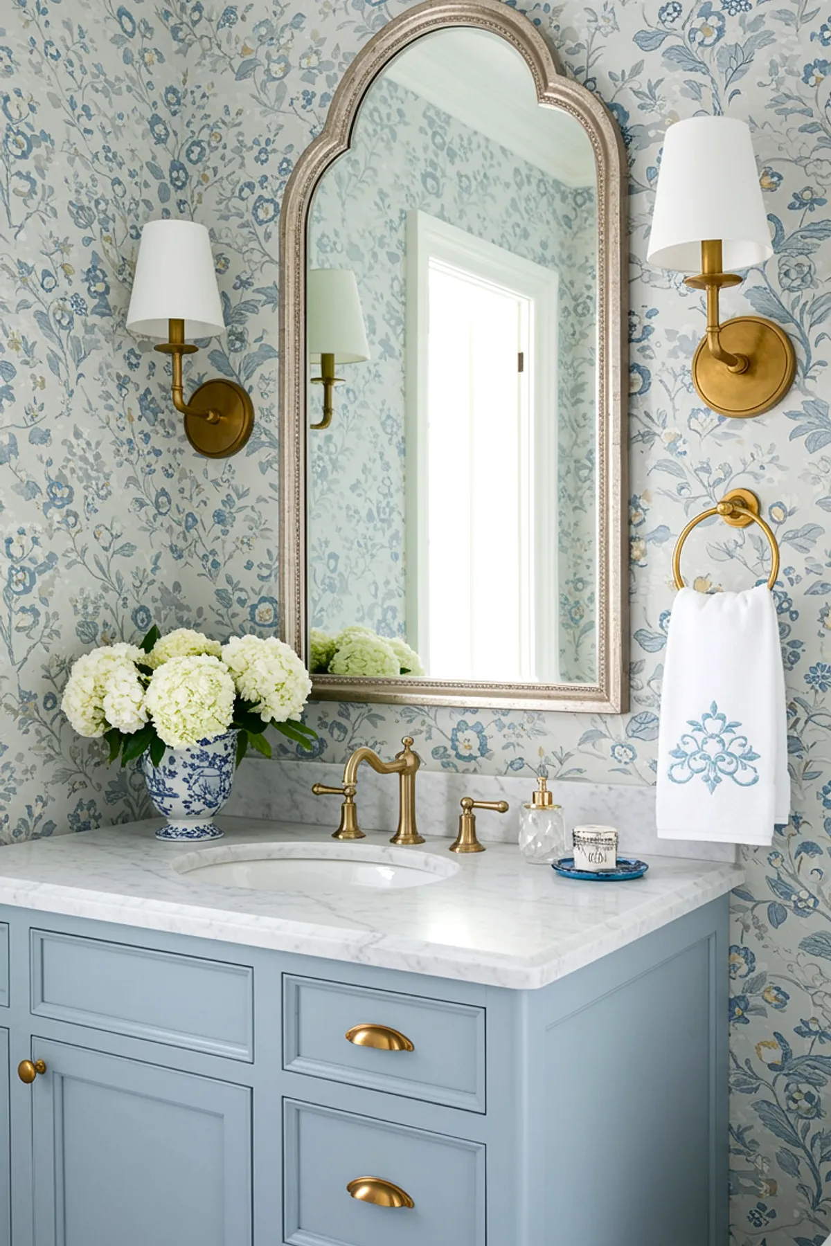 A bathroom featuring floral wallpaper, blue cabinetry, gold fixtures, and fresh flowers.