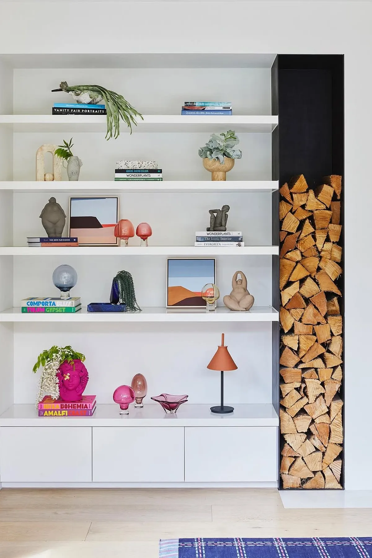 Modern shelf featuring colorful decor, sculptures, books, and a stack of firewood in a stylish living room.