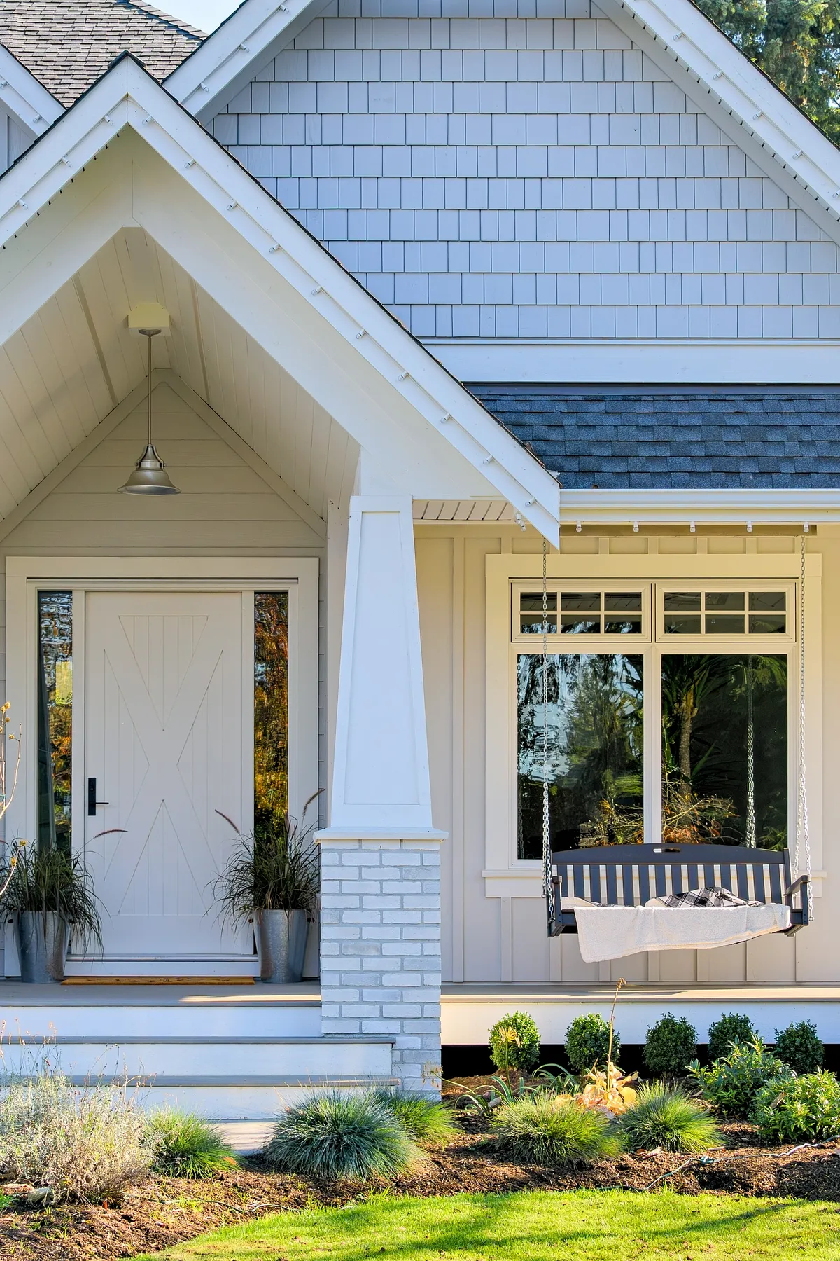 Gray shingled house with white porch swing, black door, and landscaped garden featuring lush greenery.