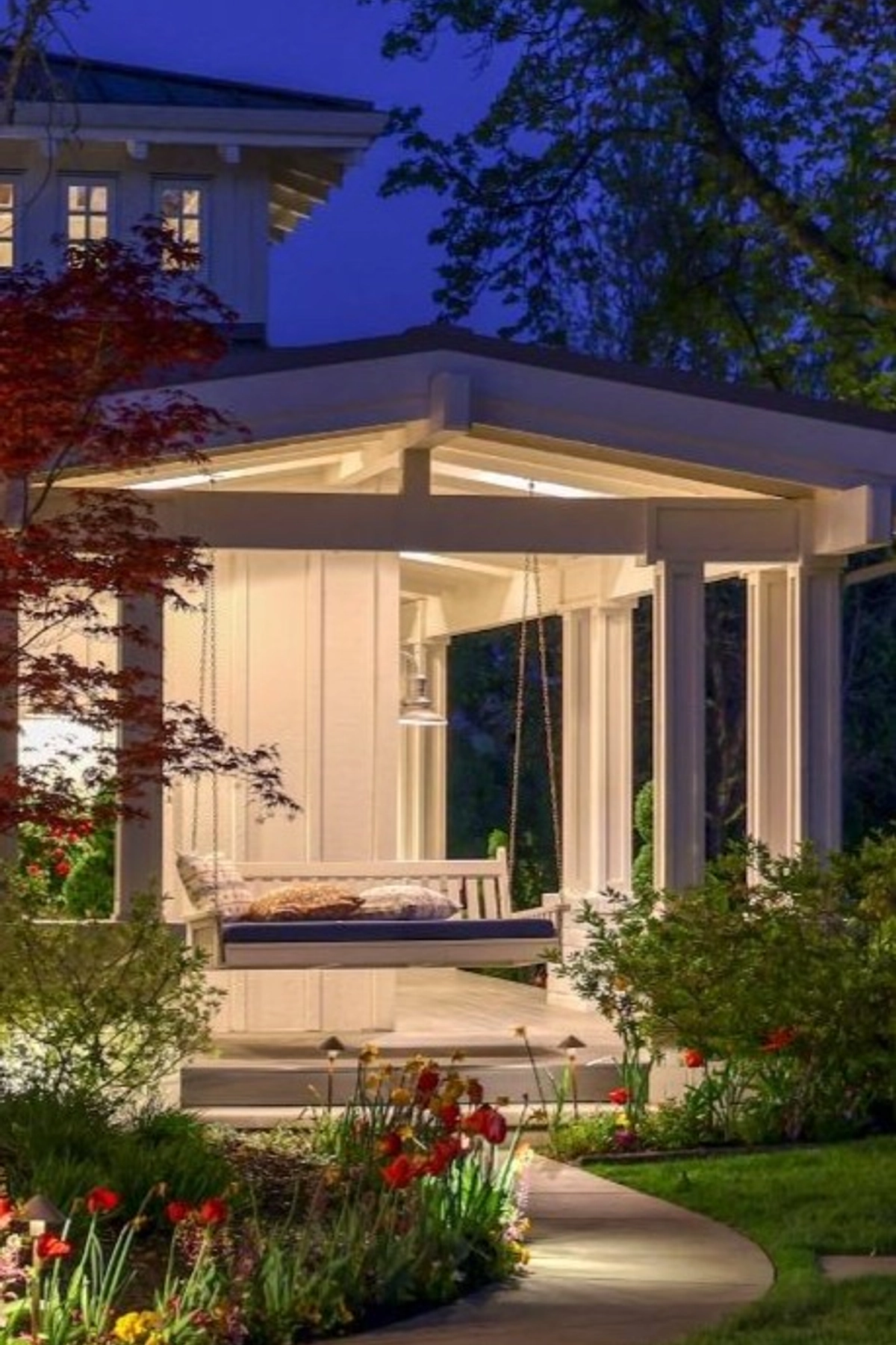 White wooden porch swing with cushions hanging from a covered pavilion, surrounded by colorful flower beds and soft evening lighting