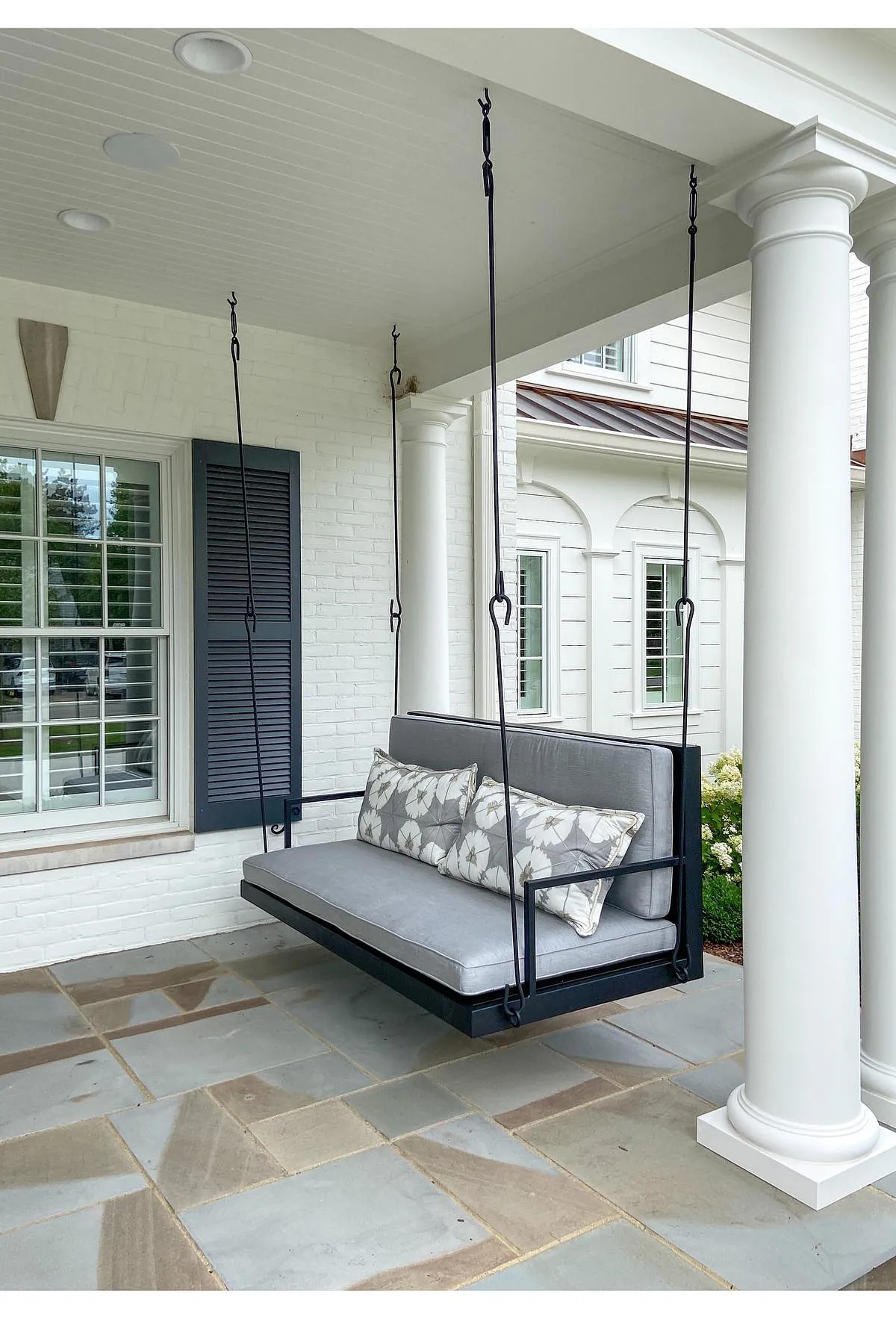 A chic porch swing in gray and black featuring floral pillows, suspended from white columns.
