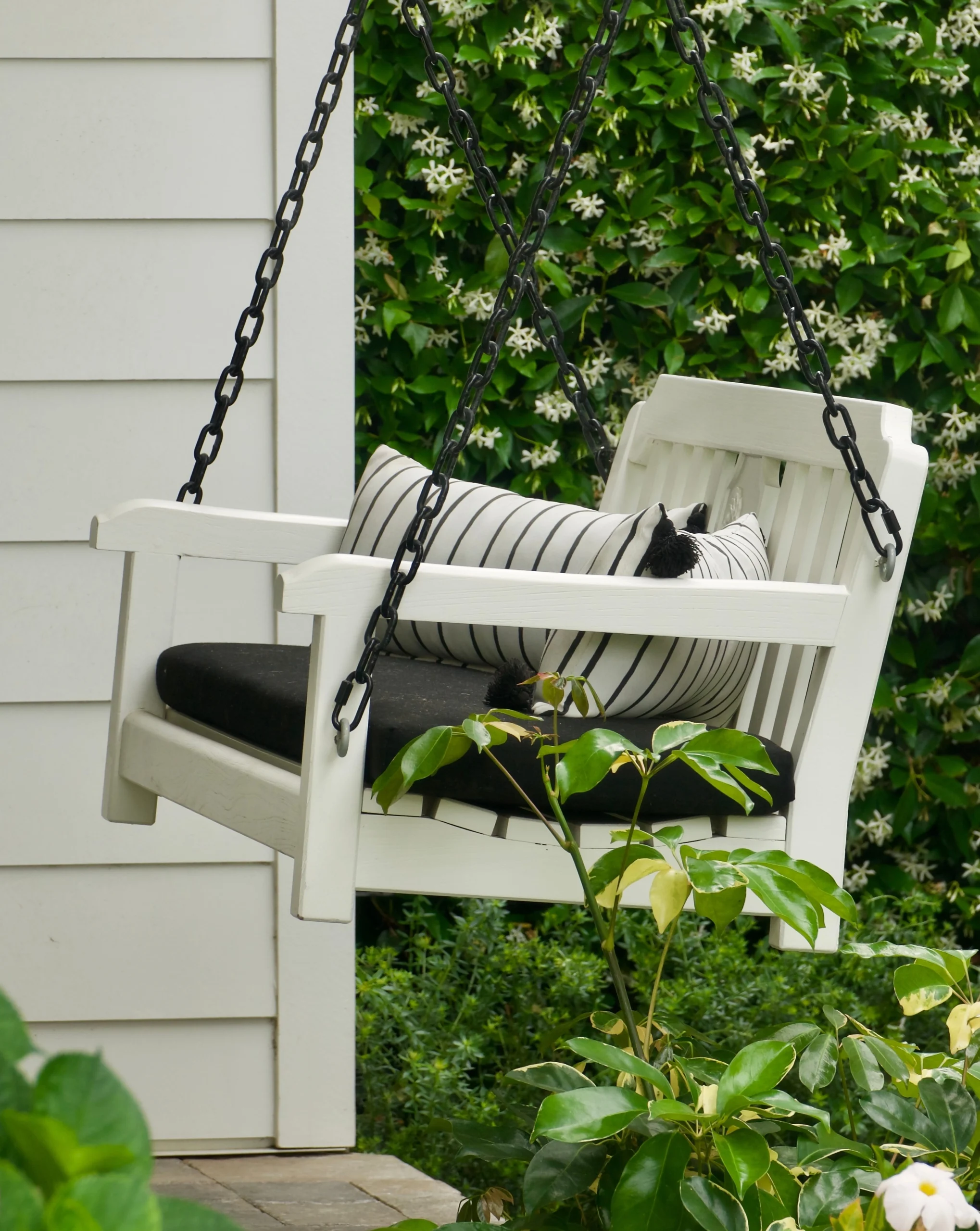 White porch swing featuring black cushion and striped pillows, set against a backdrop of greenery.