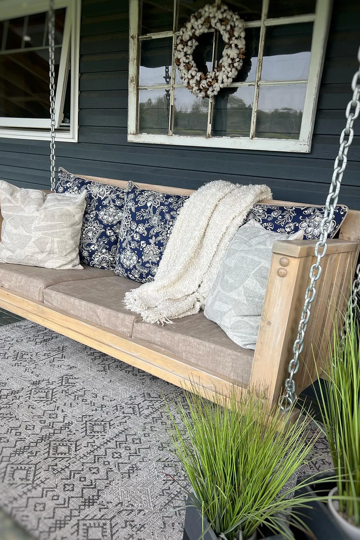 A cozy porch swing with navy and gray pillows, featuring a knitted throw and set against a vintage wreath.