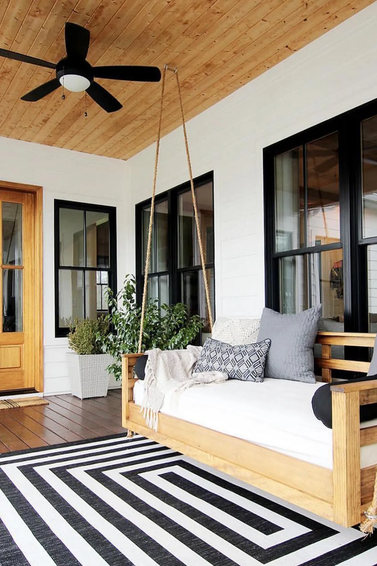 A wooden porch swing with gray pillows on a striped black and white rug, beneath a ceiling fan.