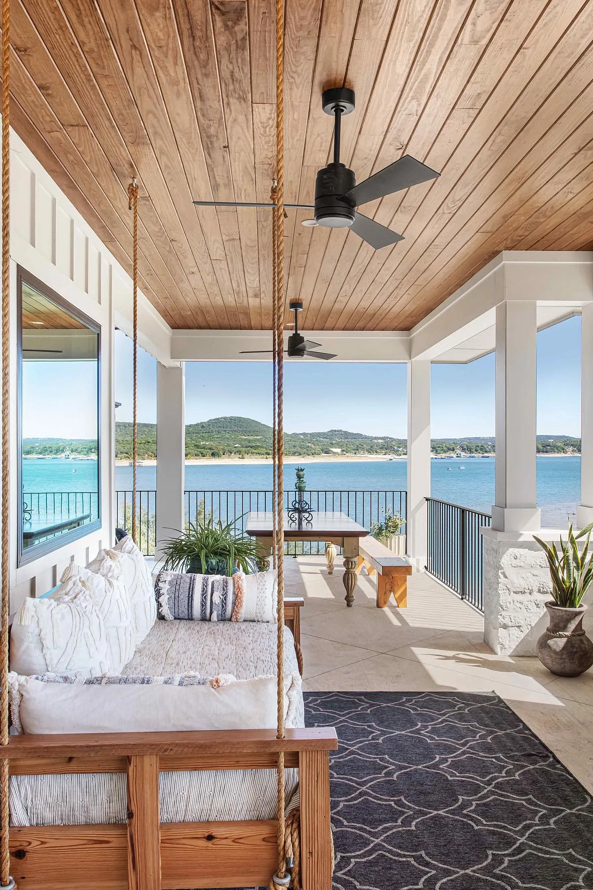 A wooden porch swing with white cushions, overlooking a serene coastal view from a covered terrace.