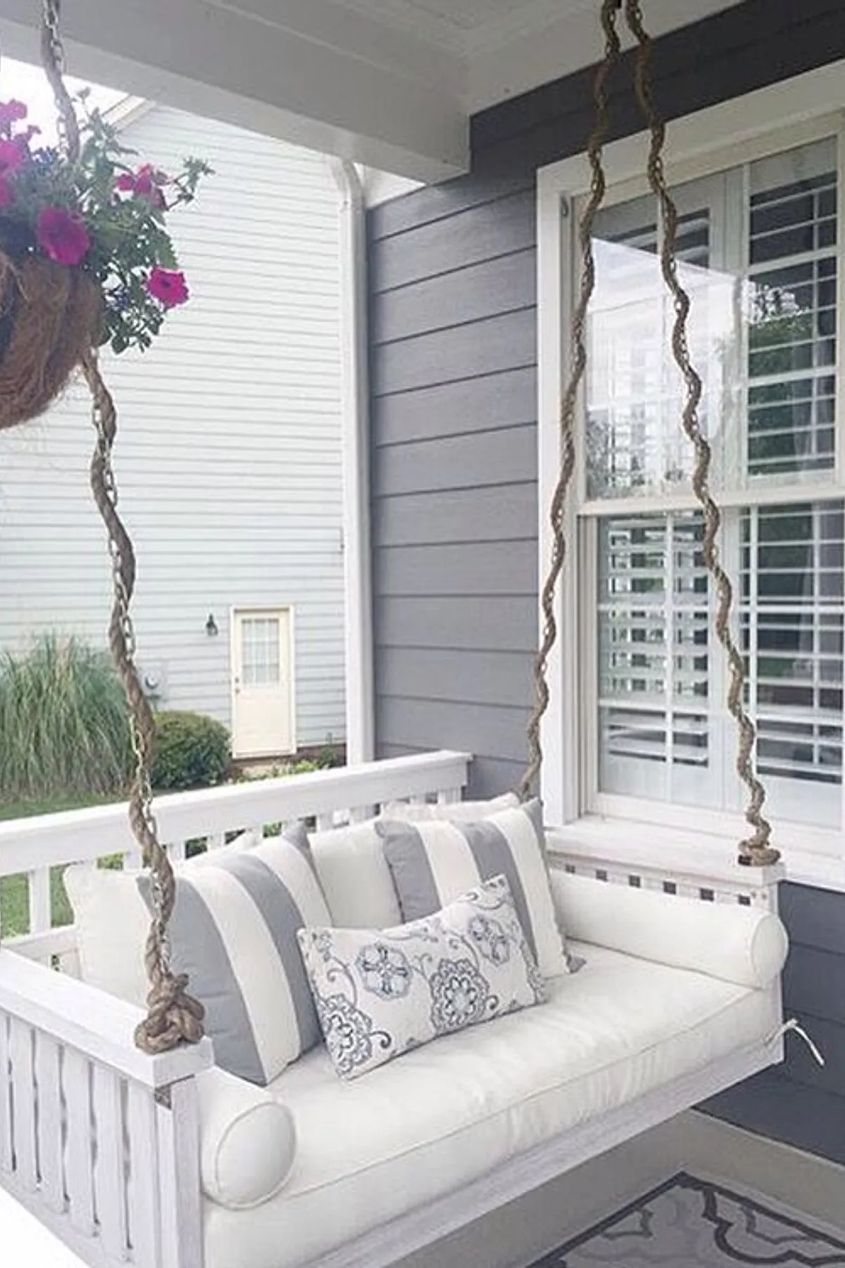 A charming porch swing with white cushions, gray striped pillows, and hanging plants against a modern backdrop.