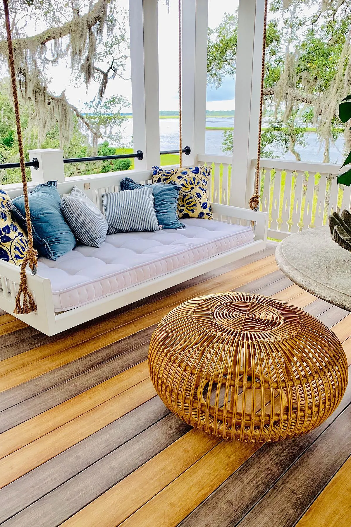 A white porch swing adorned with blue and yellow pillows, alongside a bamboo ottoman on wooden decking.