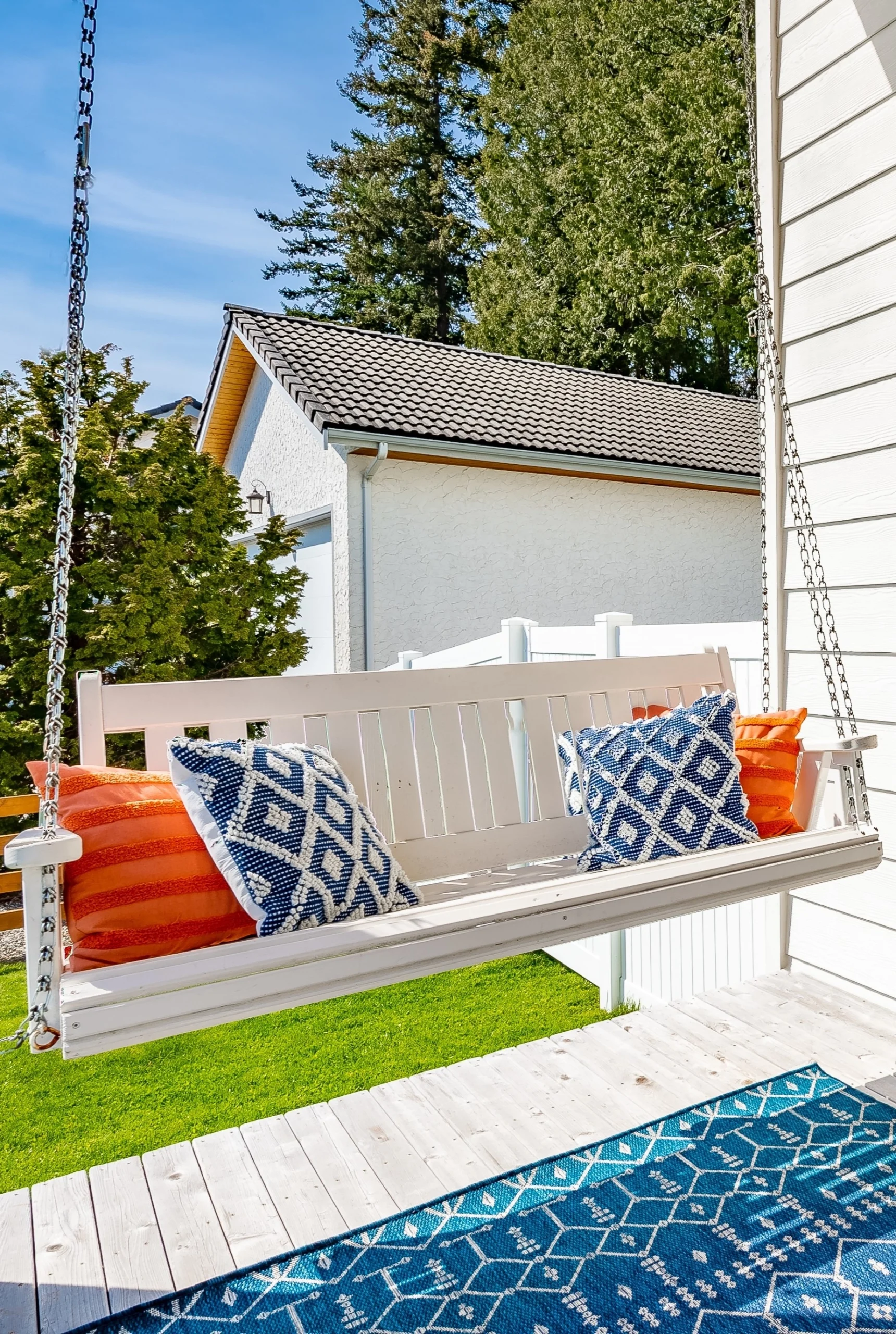 White porch swing with blue and orange pillows, set on a wooden deck with a patterned blue rug