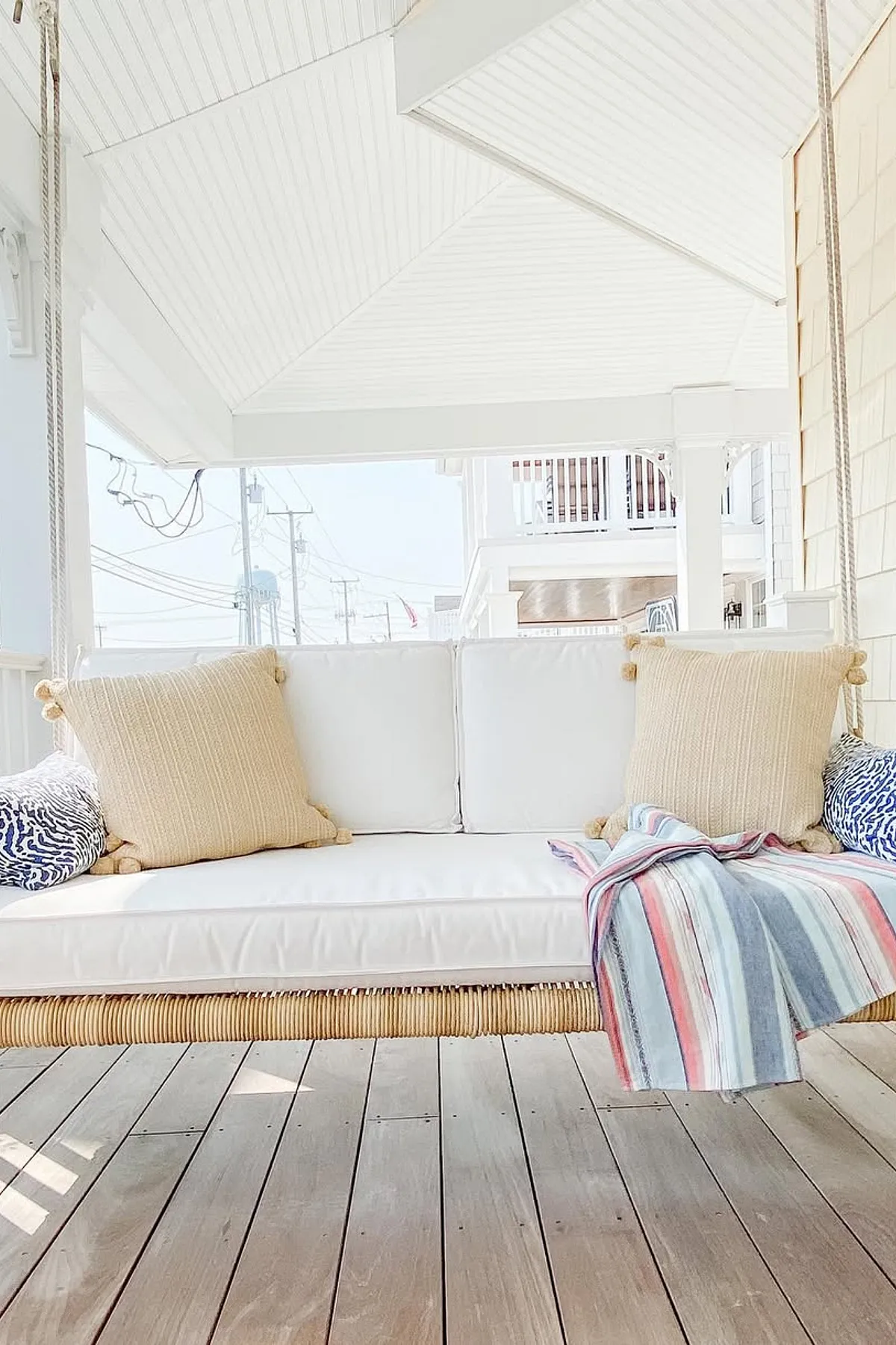 A cozy porch swing with white cushions, beige pillows, and a colorful striped throw on a wooden deck.