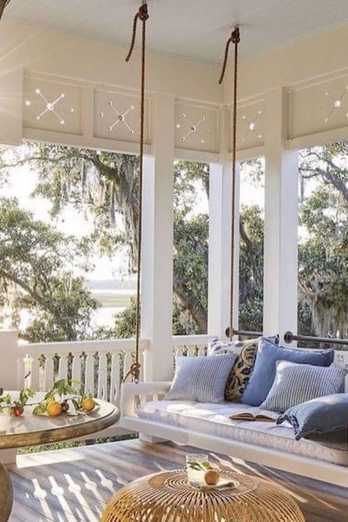 A white porch swing with blue striped pillows, suspended in a bright space with natural wood flooring.