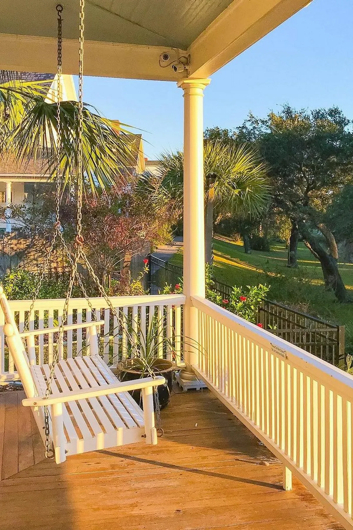 A white porch swing on a sunny deck surrounded by potted plants and vibrant garden views.