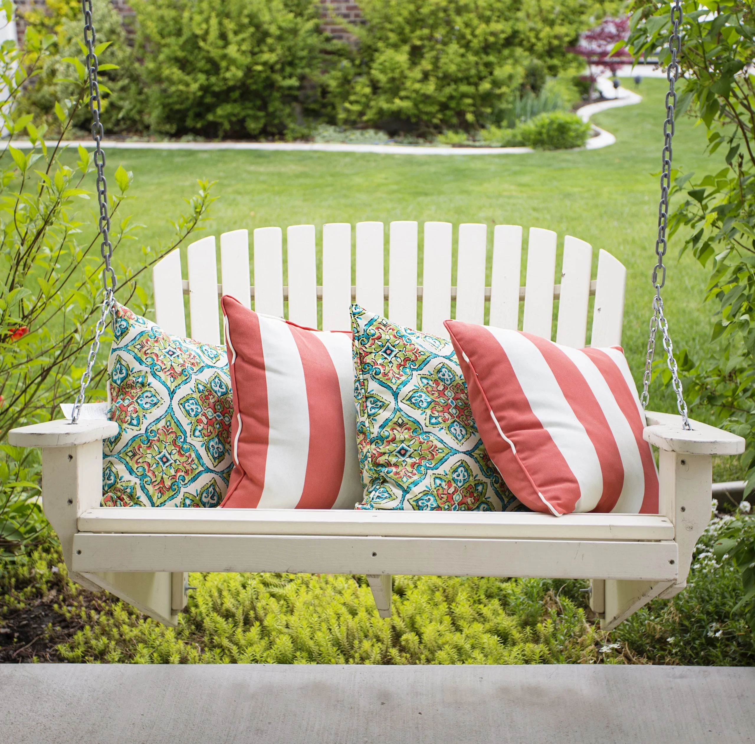 White porch swing featuring colorful coral and patterned pillows, set against a backdrop of green foliage.