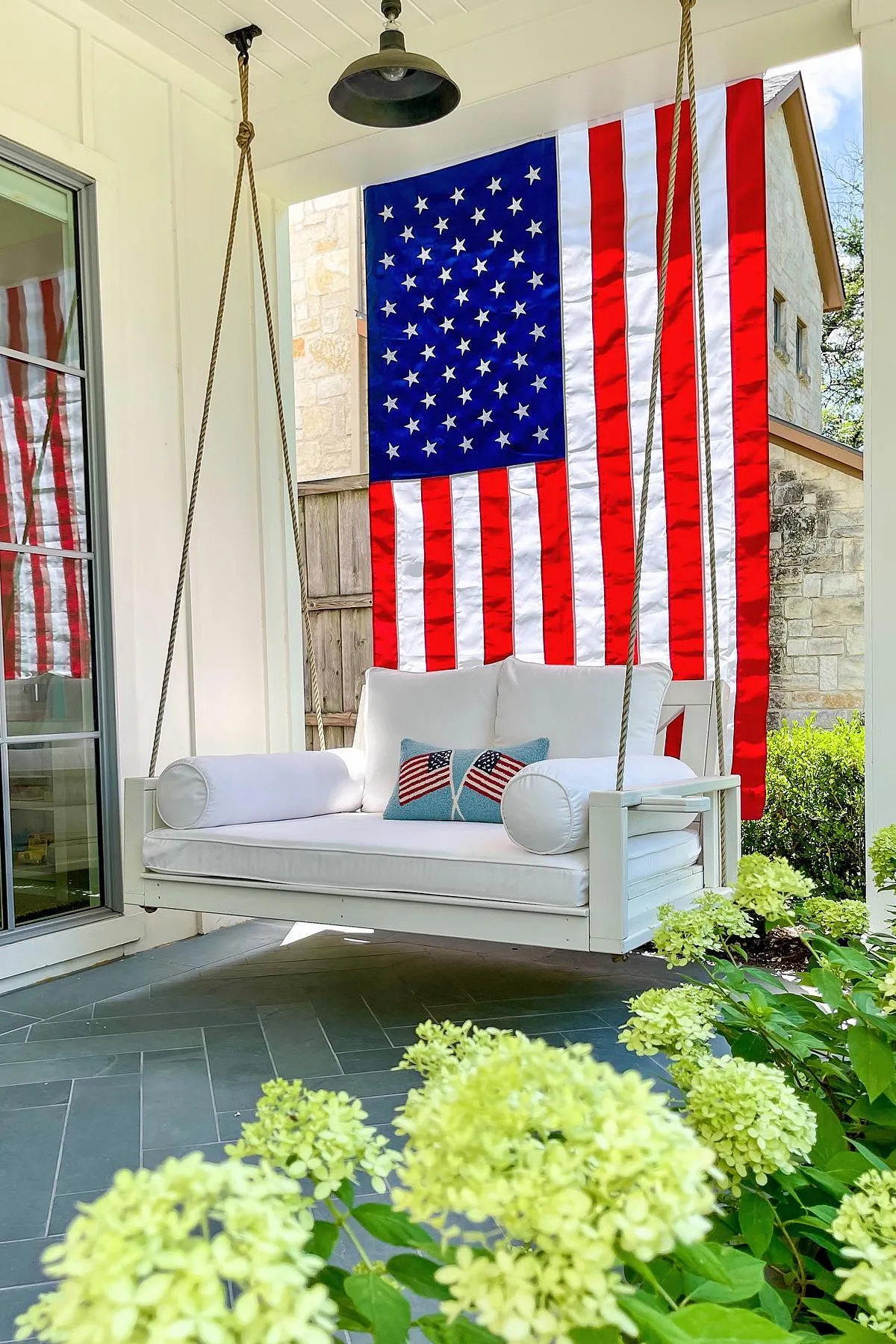 A cozy porch swing adorned with a decorative American flag and pillows, surrounded by lush greenery.