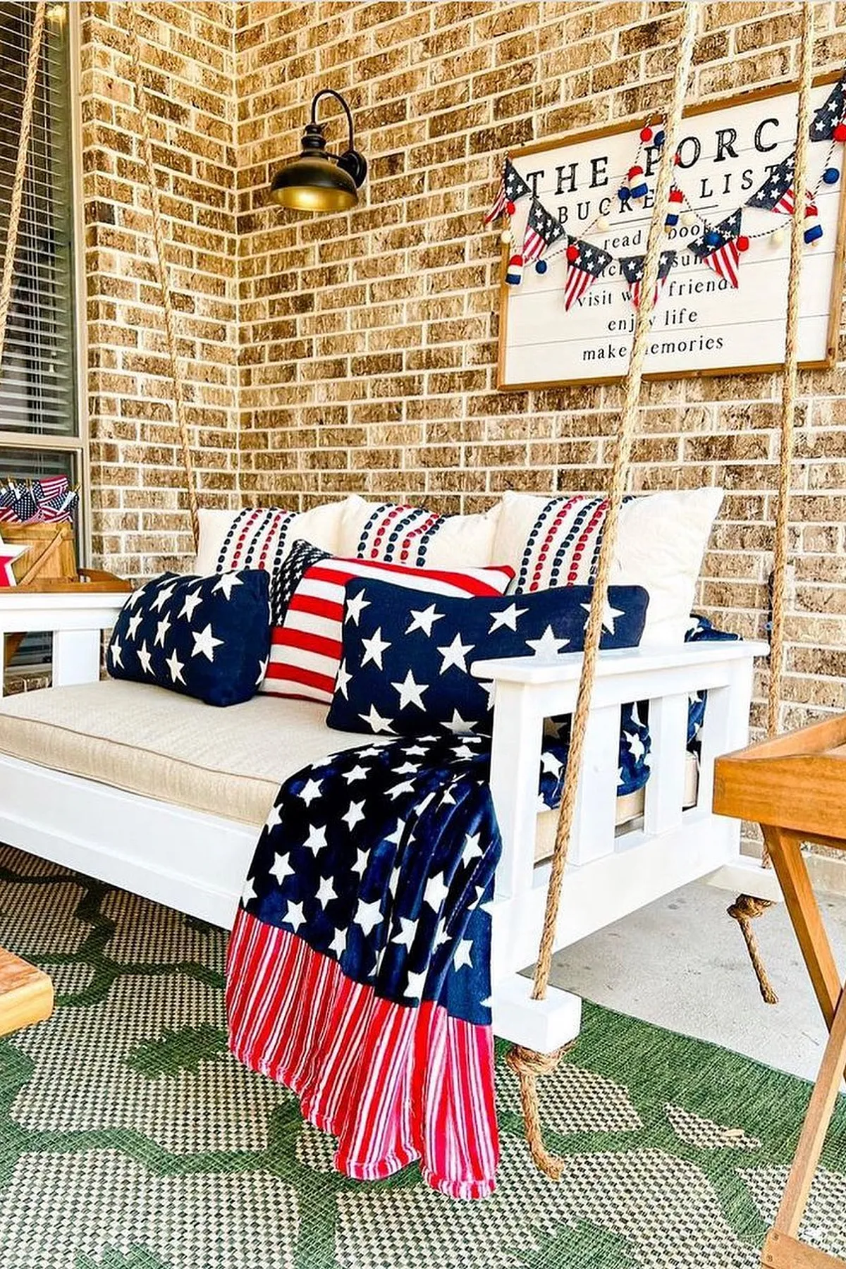 A porch swing decorated with star-patterned pillows and blankets, alongside a rustic sign and greenery.