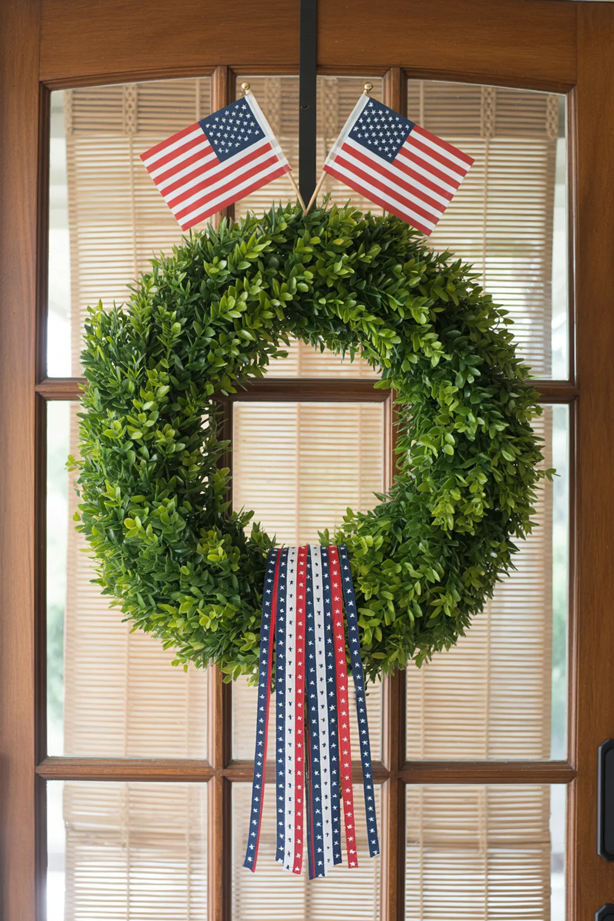 A green wreath adorned with American flags and ribbons in patriotic colors, hanging on a wooden door.