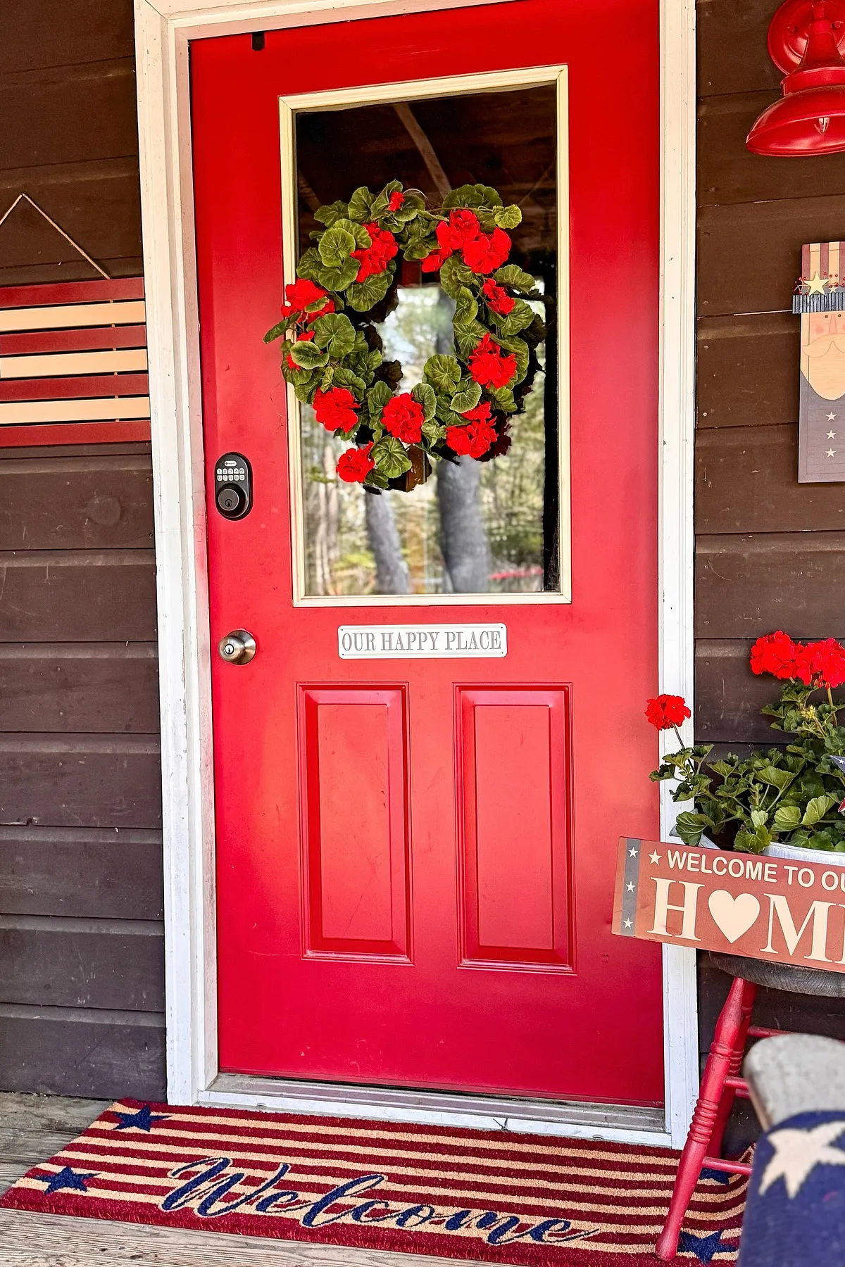 A bright red door with a floral wreath, 'Our Happy Place' sign, and patriotic welcome mat on a cozy porch.