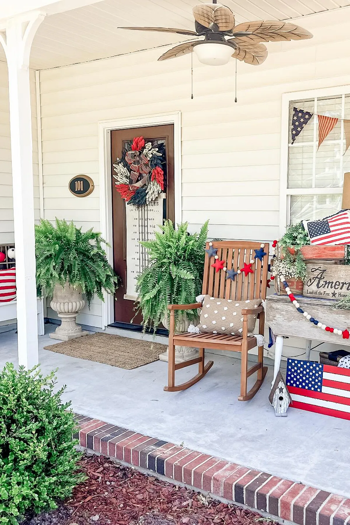 A festive porch featuring a patriotic wreath, potted ferns, rocking chair with decorative pillow, and colorful decor.