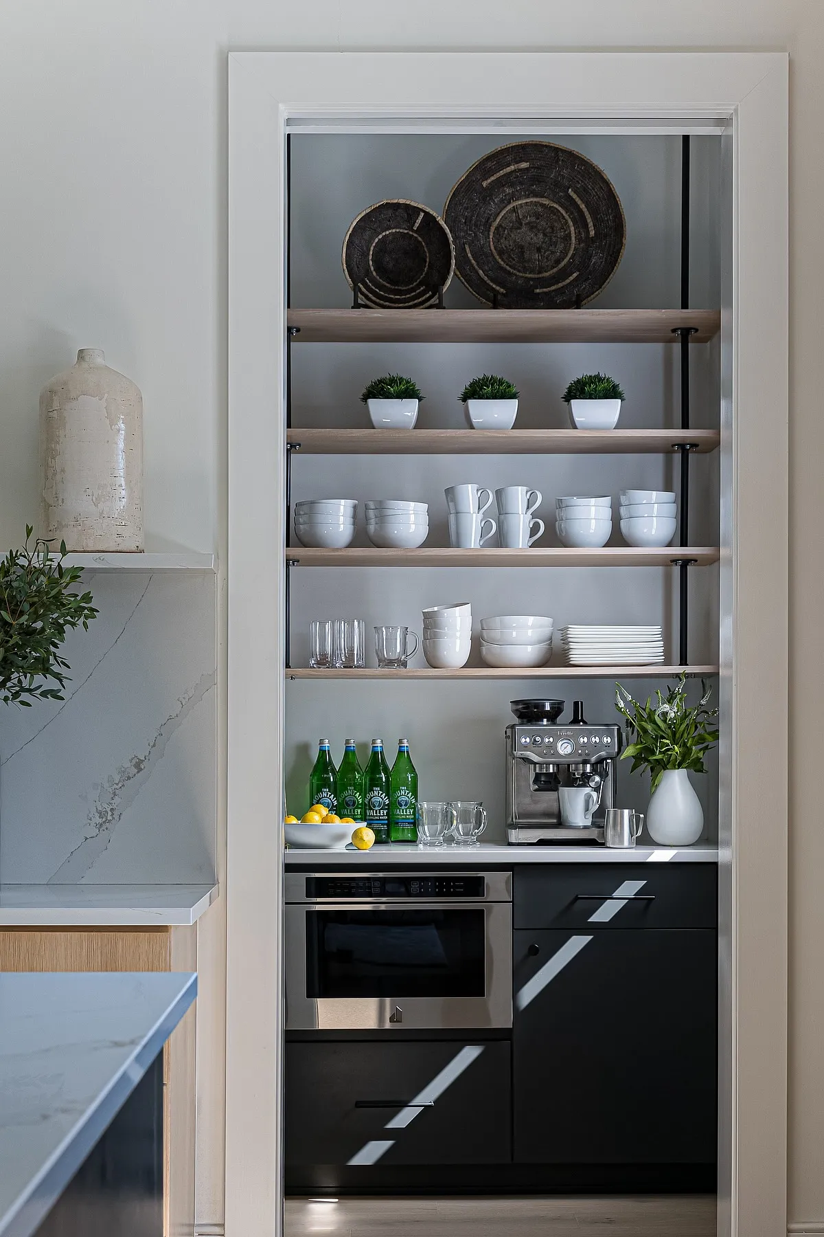 Coffee station with multi-level shelves holding white dishes, mugs, small plants, and an espresso machine.