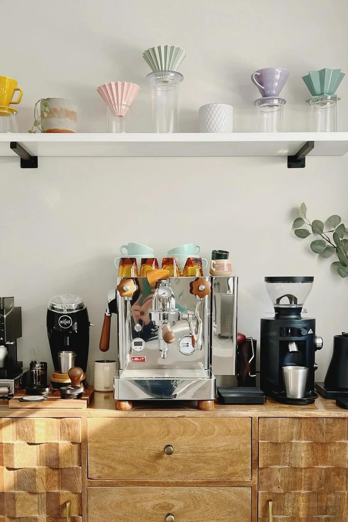Coffee station with wooden cabinet, espresso machine, and colorful ceramic mugs on a white shelf.