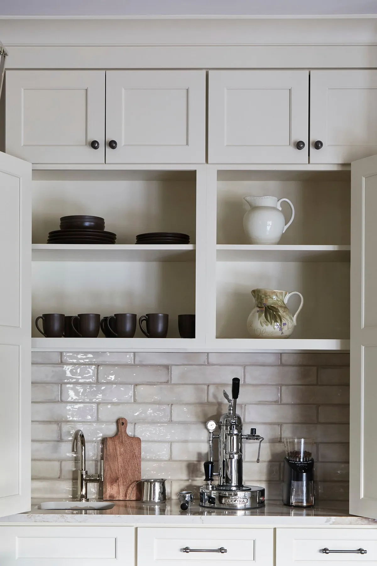 Coffee station with white cabinets, dark mugs, glossy subway tile backsplash, and silver espresso machine.