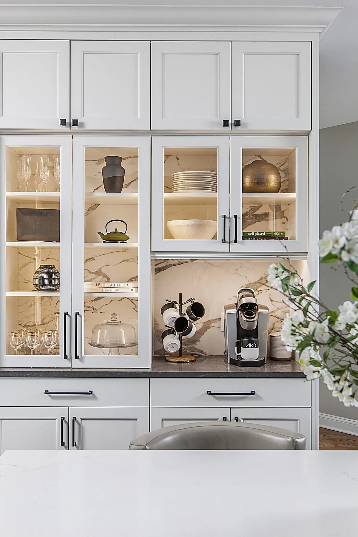 White kitchen cabinets with glass fronts displaying ceramics and a coffee machine on the counter.